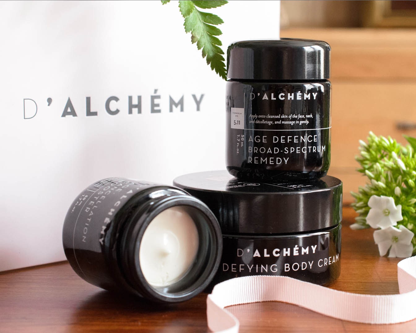 Na zdjęciu znajdują się trzy czarne słoiczki kosmetyków D'ALCHÉMY: Age Defence Broad-Spectrum Remedy oraz Defying Body Cream, ułożone na drewnianej powierzchni, obok białych kwiatów i wstążki.