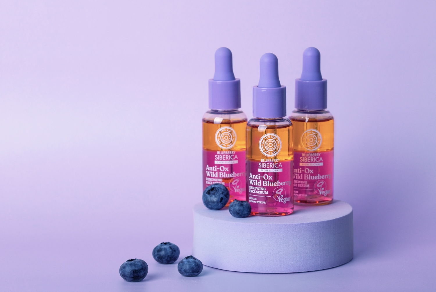 Trois flacons de sérum pour le visage Blueberry Siberica Anti-Ox Wild Blueberry, avec un liquide doré et un emballage rose et violet, sont présentés avec des myrtilles fraîches.