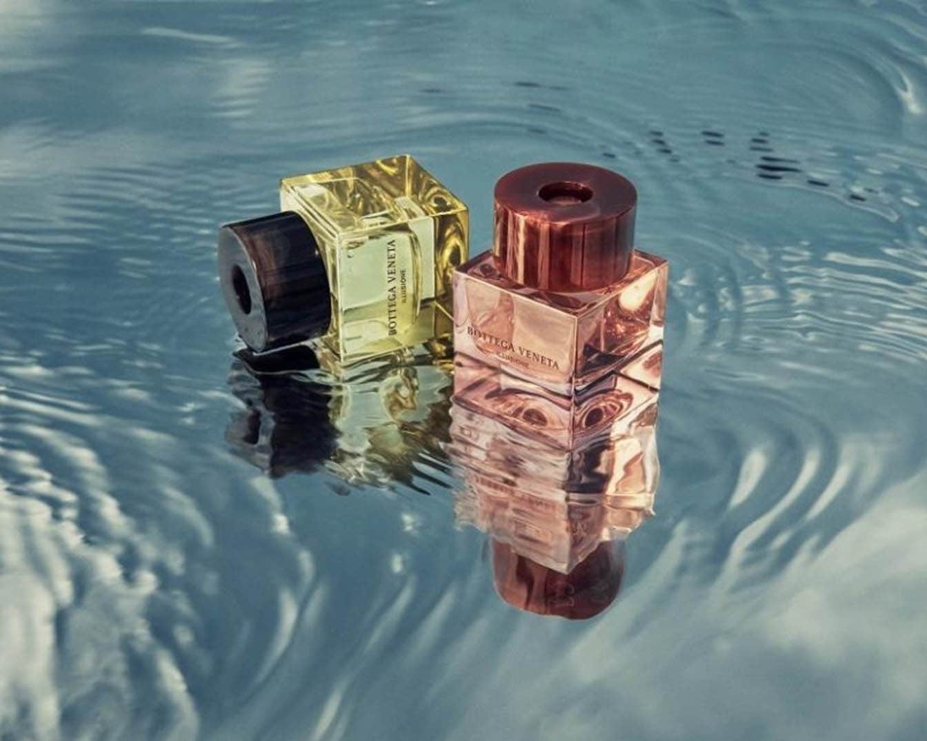Dwa flakony perfum Bottega Veneta Illusione, jeden złoty z ciemnobrązową nakrętką, drugi różowy z miedzianą nakrętką, stoją na powierzchni wody, tworząc odbicie.