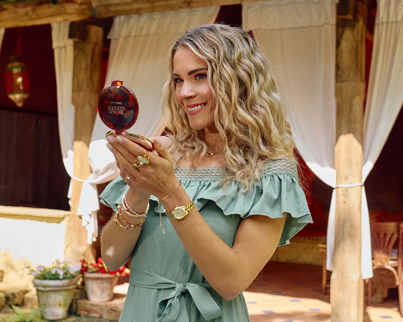 Une femme souriante, vêtue d'une robe vert d'eau, tient un poudrier Bronze Goddess d'Estée Lauder, reflétant un teint radieux et ensoleillé.