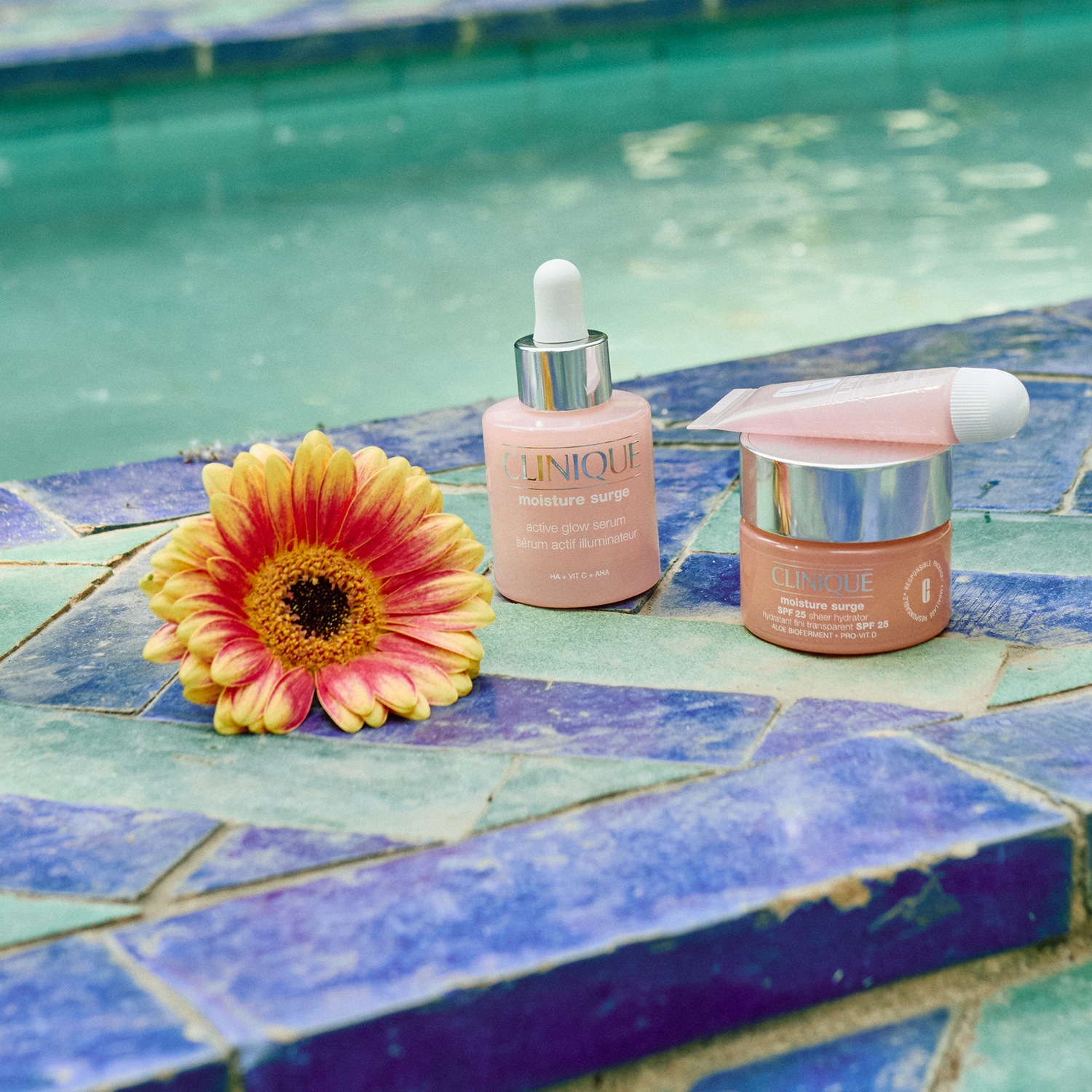 Sur une mosaïque bleue et verte, découvrez le sérum éclat actif Moisture Surge de Clinique dans son flacon rose, accompagné d'une fleur jaune et orange.
