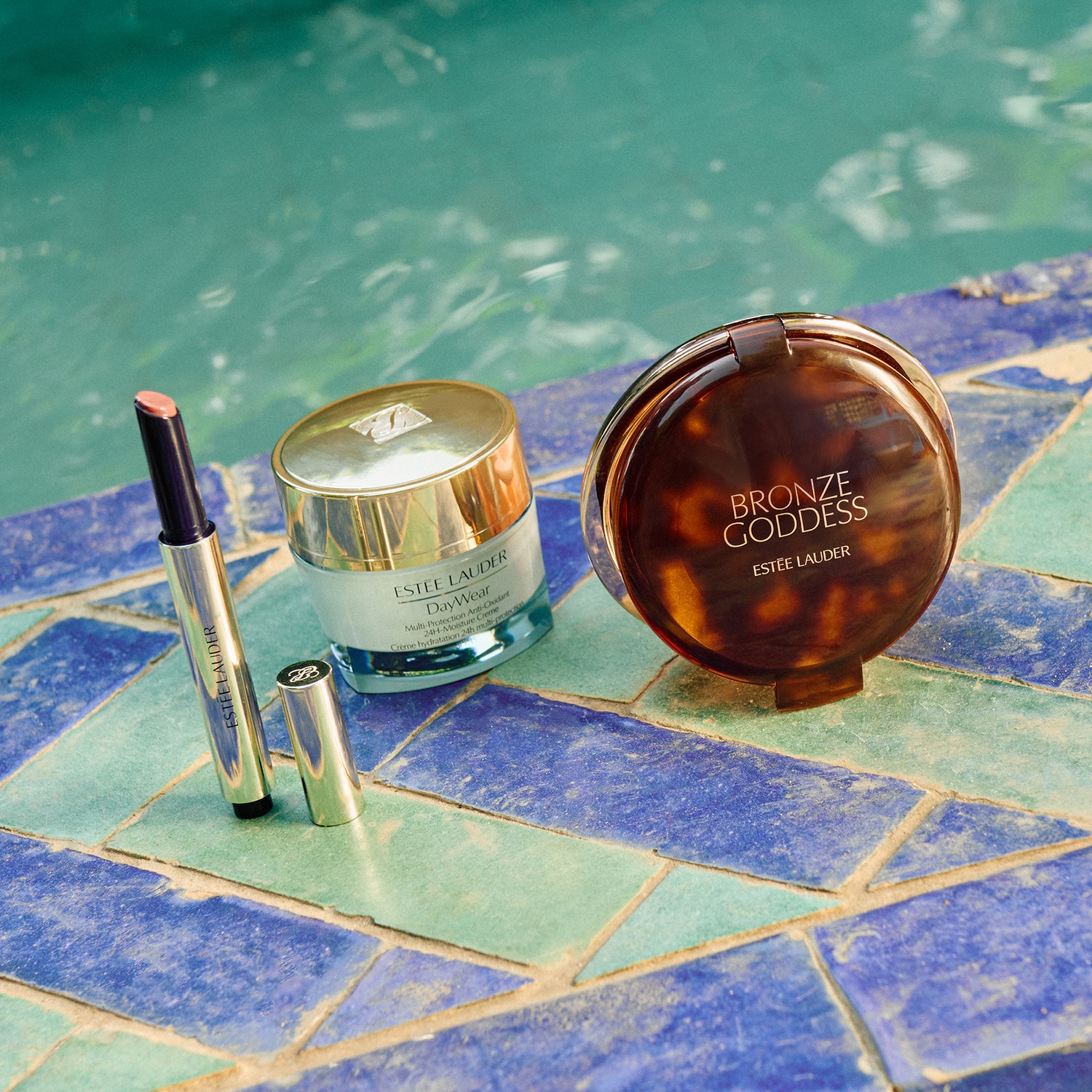 Découvrez le rouge à lèvres Estée Lauder, le soin hydratant DayWear et la poudre bronzante Bronze Goddess, présentés sur un fond de mosaïque bleue et verte.
