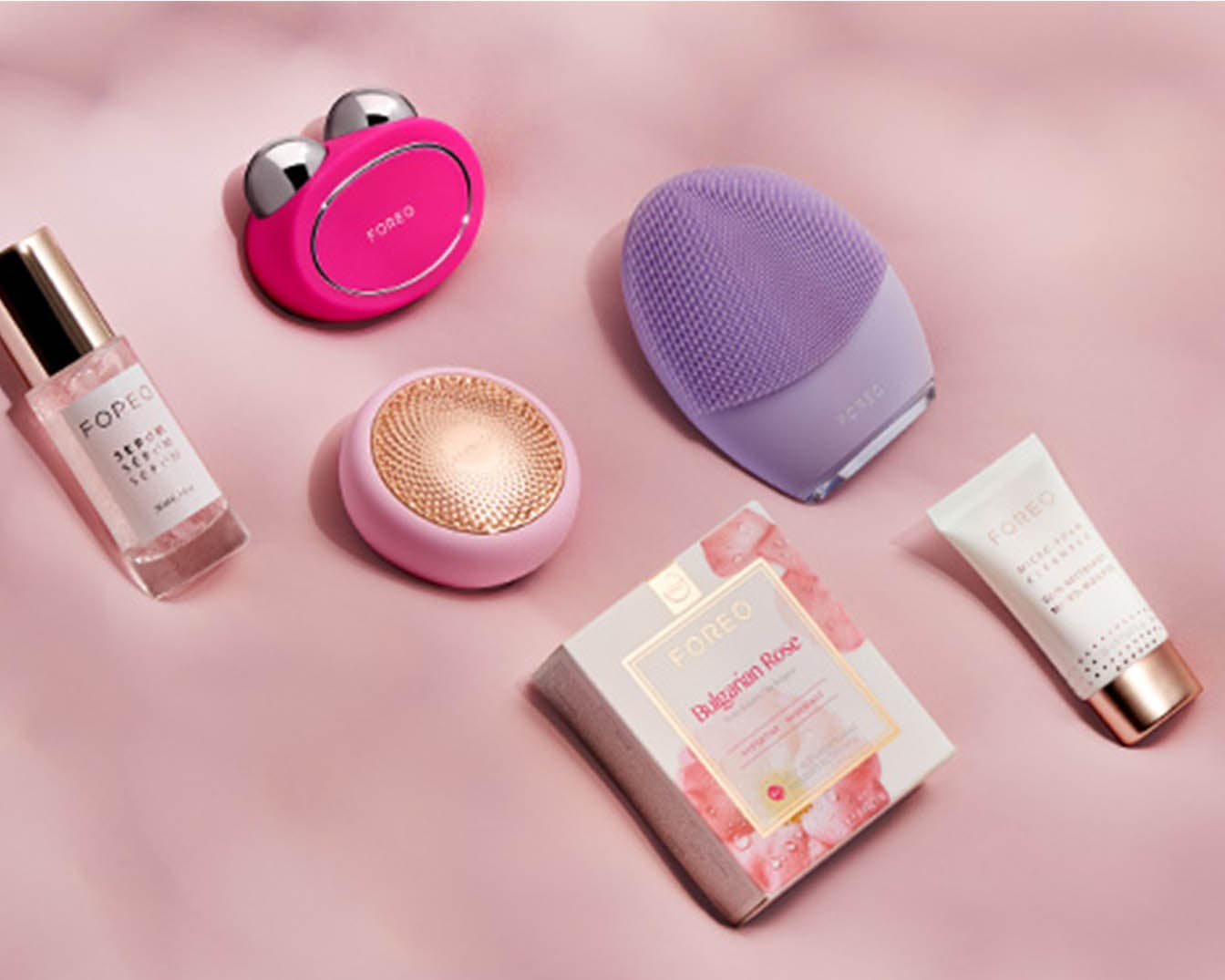 Różowa szczoteczka do twarzy Foreo, fioletowa szczoteczka, serum w przezroczystej butelce, krem w białej tubce i pudełko Bulgarian Rose na różowym tle.