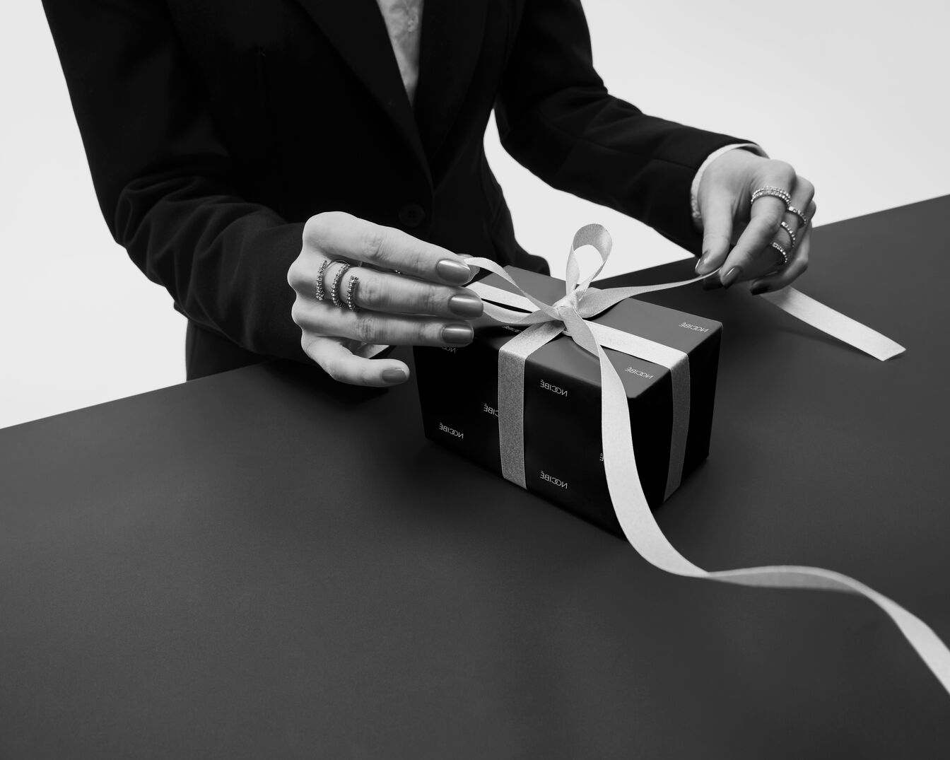 Une personne en costume noir ouvre un cadeau emballé dans du papier noir orné d'un ruban blanc, ajoutant une touche d'élégance à ce moment spécial.