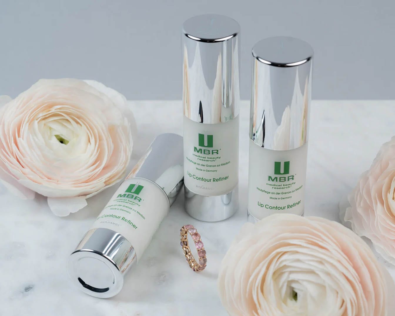 Trzy białe butelki MBR Lip Contour Refiner z srebrnymi nakrętkami stoją obok różowych kwiatów i pierścionka, tworząc elegancką kompozycję.