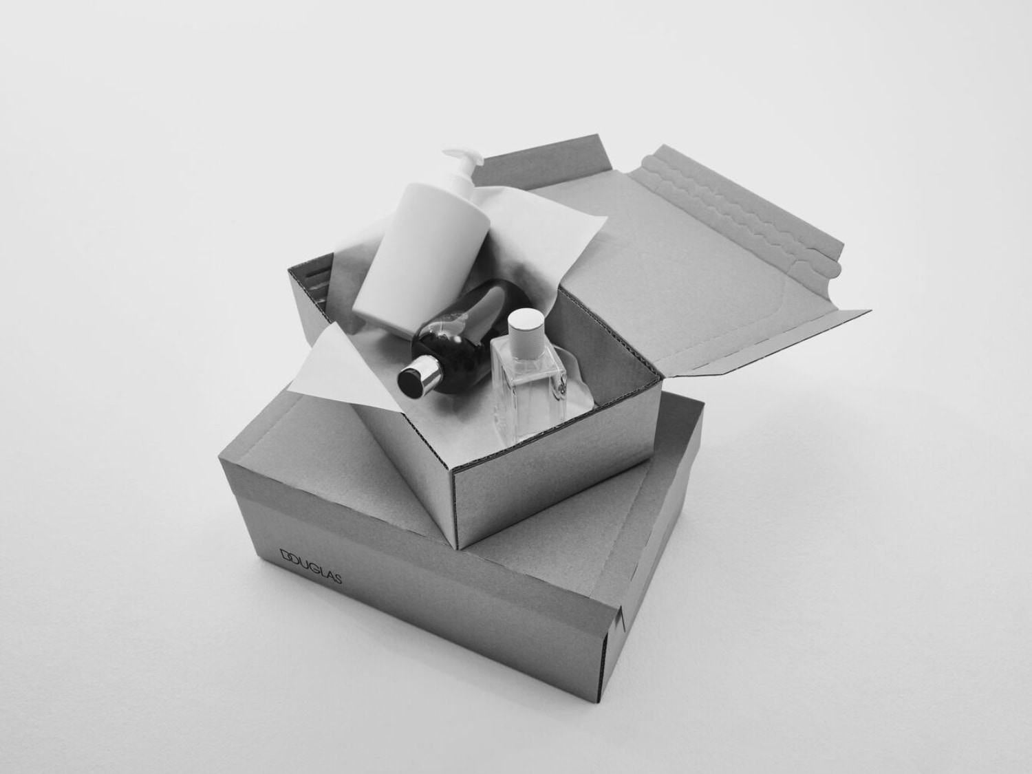 Na zdjęciu widać otwarte kartonowe pudełko Douglas, w którym znajdują się biała butelka z pompką, ciemna butelka i przezroczysty flakon perfum.