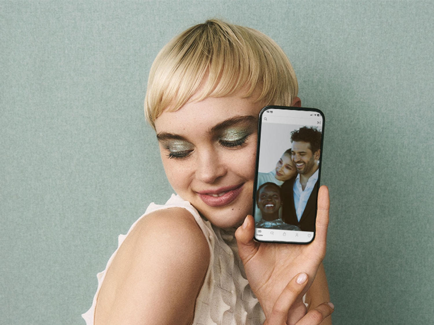 Kobieta z krótkimi blond włosami i zielonym makijażem oczu uśmiecha się, trzymając telefon, na którego ekranie widać zdjęcie trzech uśmiechniętych osób.