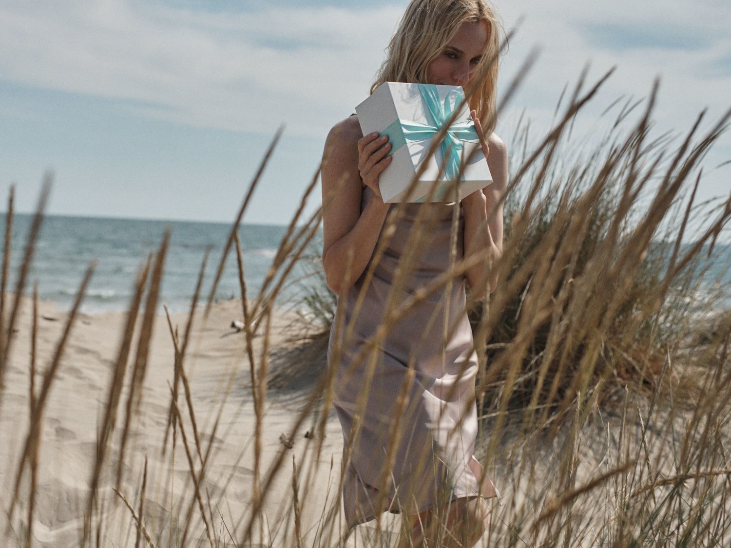 Sur une plage ensoleillée, une femme blonde, vêtue d'une robe légère, tient un cadeau blanc orné d'un ruban turquoise, entourée d'herbes sauvages.