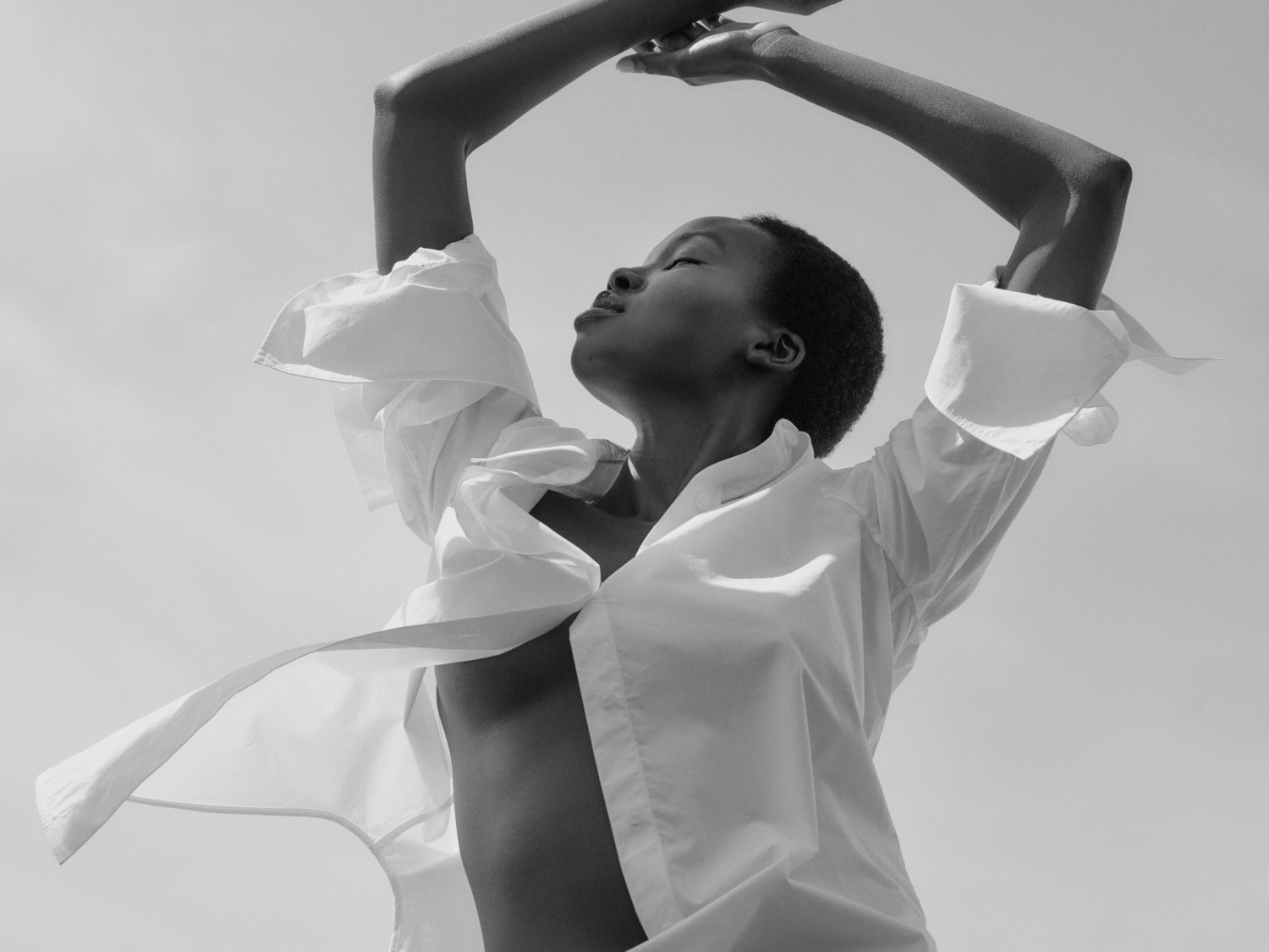 En contre-plongée, une femme à la peau foncée, vêtue d'une chemise blanche ouverte, lève les bras vers le ciel dans un geste de liberté et de bien-être.