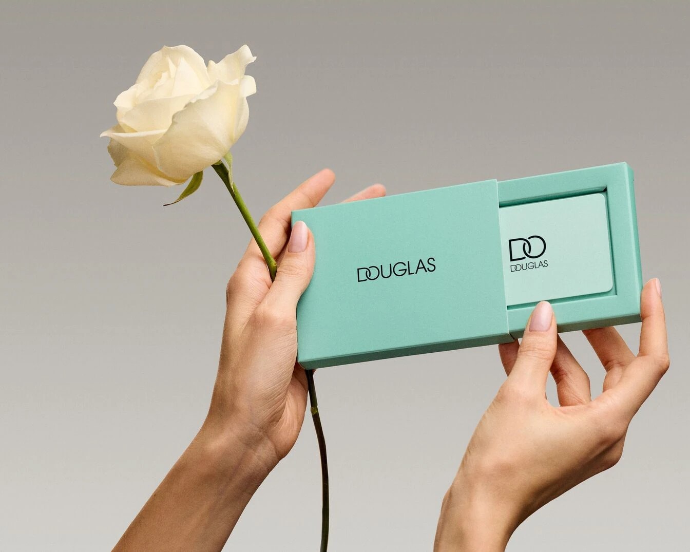 Dłonie otwierają turkusowe pudełko Douglas, w którym znajduje się karta podarunkowa, obok leży biała róża.
