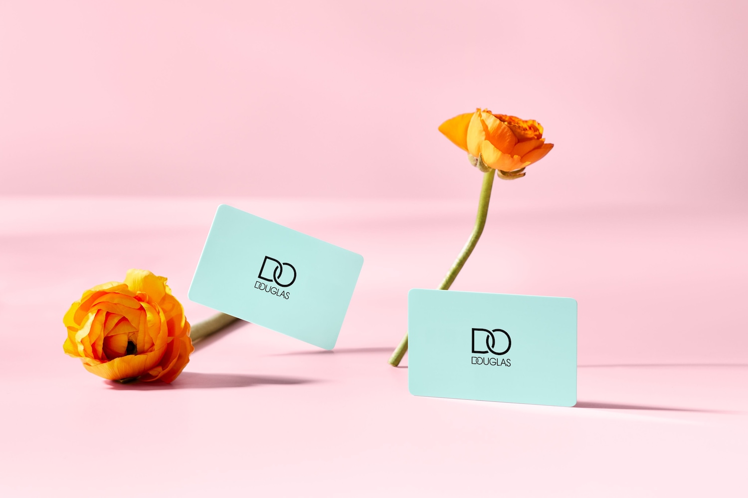 Na różowym tle leżą dwie karty podarunkowe Douglas w kolorze miętowym, obok których znajdują się pomarańczowe kwiaty.