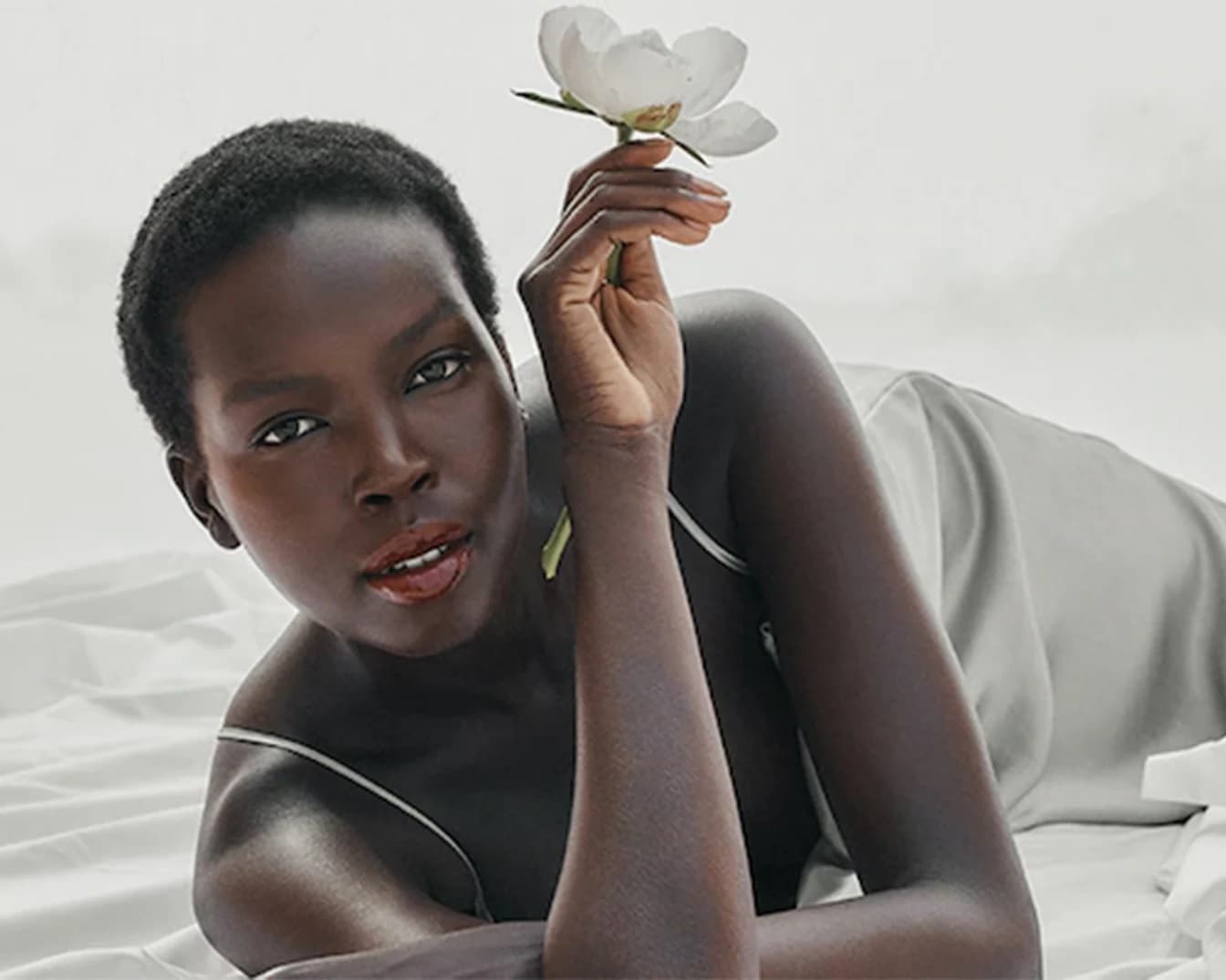 Una donna dalla pelle scura, distesa su un lenzuolo bianco, tiene delicatamente un fiore bianco tra le dita, creando un'immagine di serena bellezza.