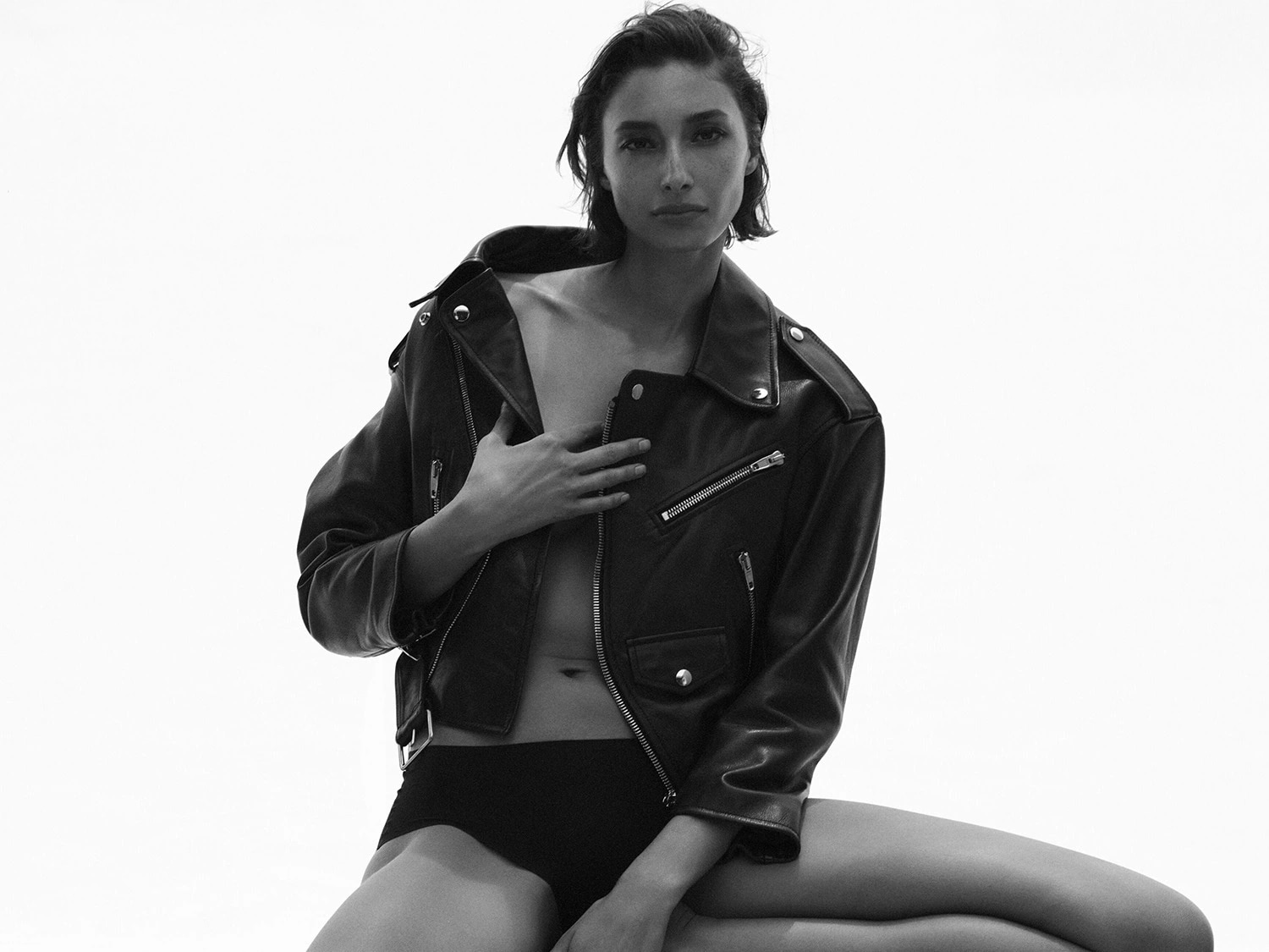 In un ritratto in bianco e nero, una modella indossa una giacca di pelle nera aperta sopra un top nero, con una mano appoggiata sul petto.