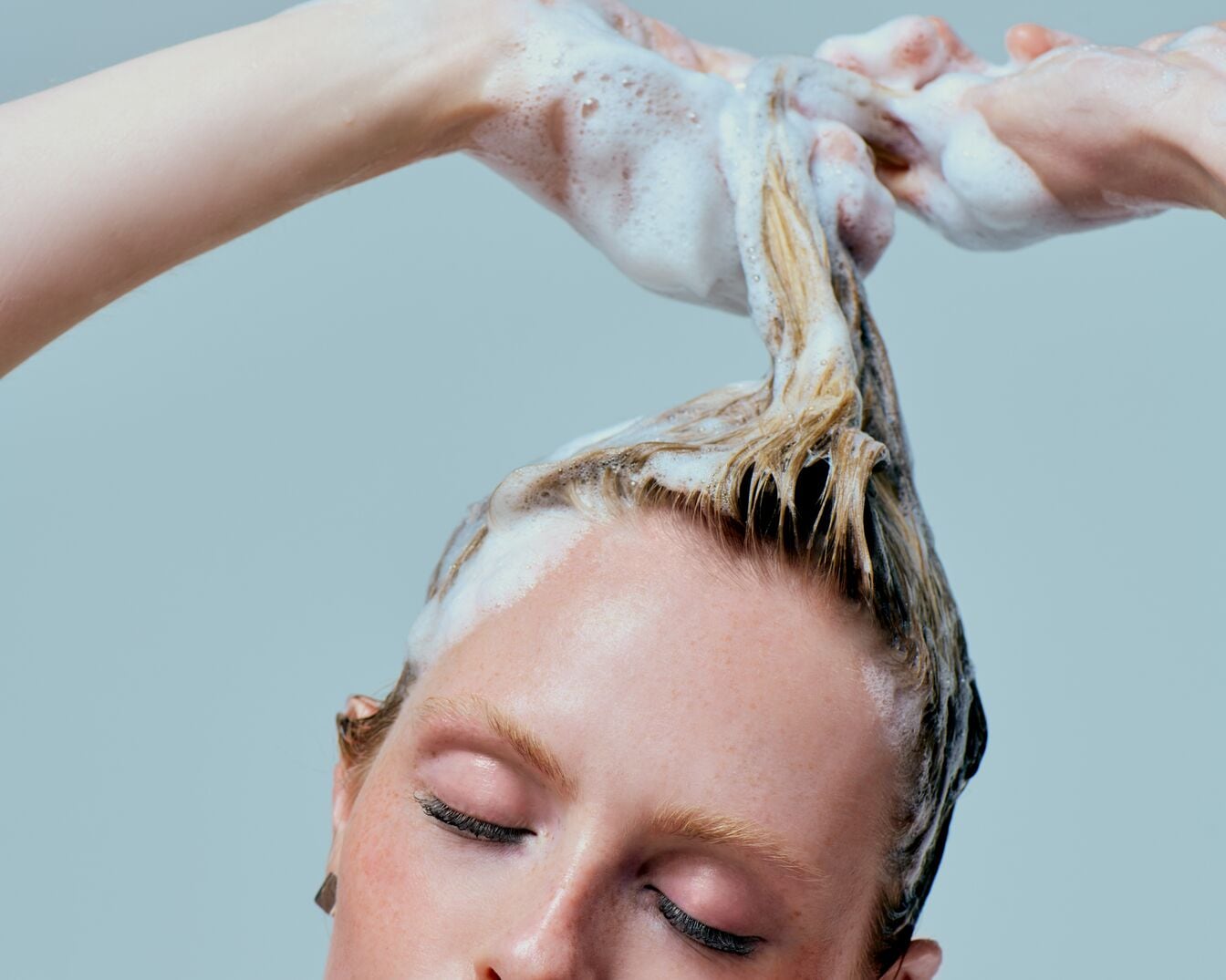 Een vrouw met gesloten ogen wast haar blonde haar met schuimende shampoo.