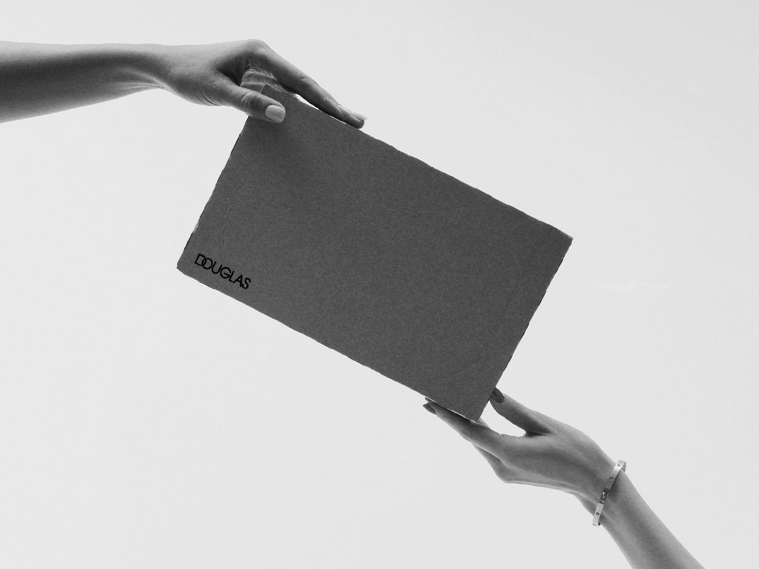 Dwie dłonie przekazują sobie szare pudełko z logo Douglas, prezentując elegancki i subtelny przekaz.