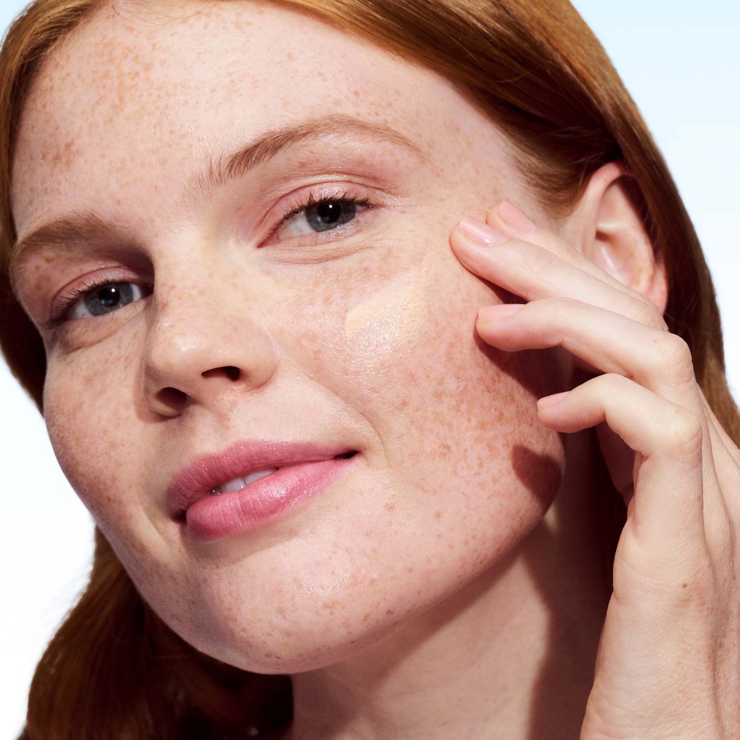 Une femme rousse applique du fond de teint beige sur sa joue, estompant doucement avec ses doigts pour un teint uniforme.