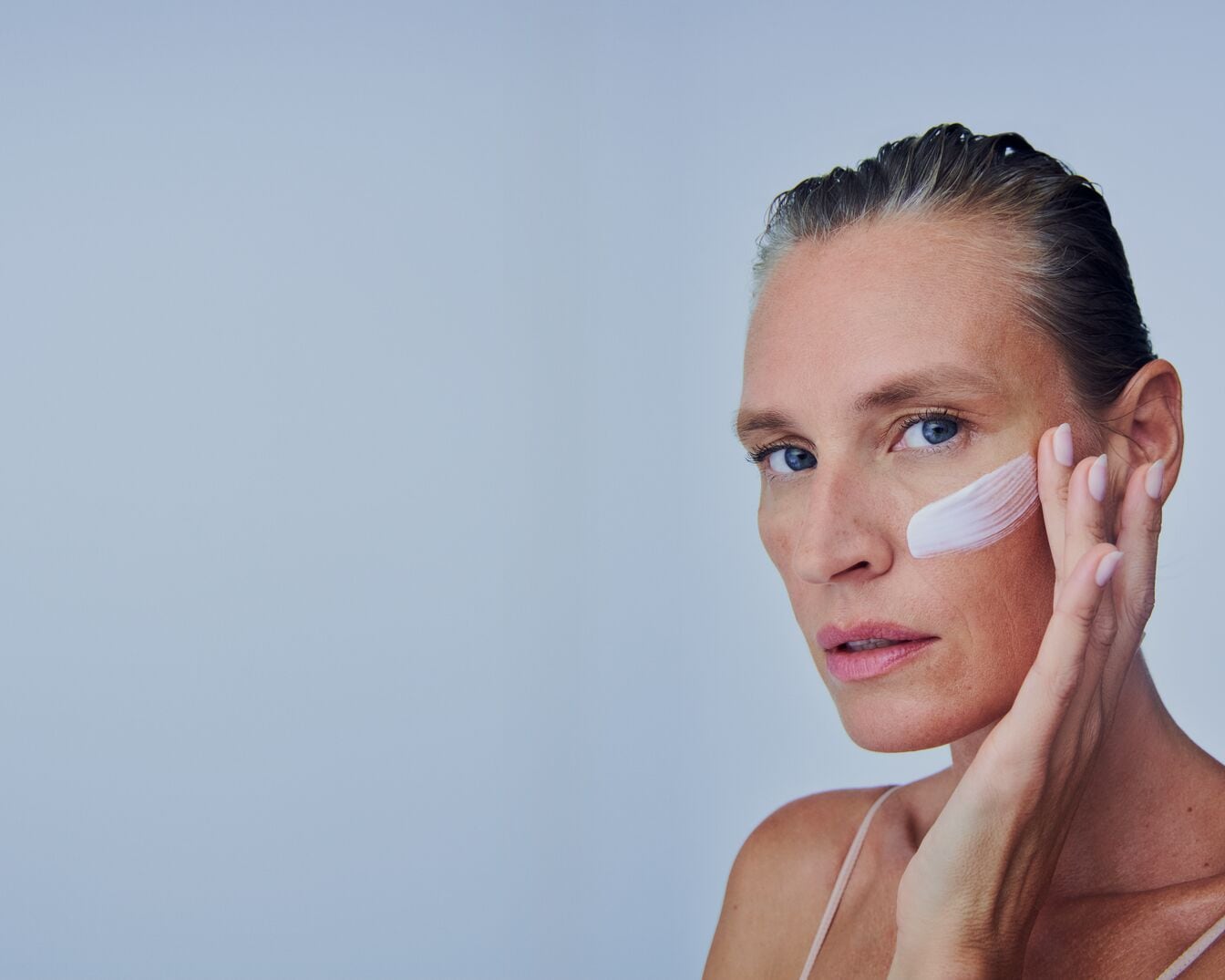 Een vrouw met blauwe ogen brengt een witte crème aan op haar gezicht, waarbij ze haar vingers gebruikt om de crème zachtjes in haar huid te masseren.