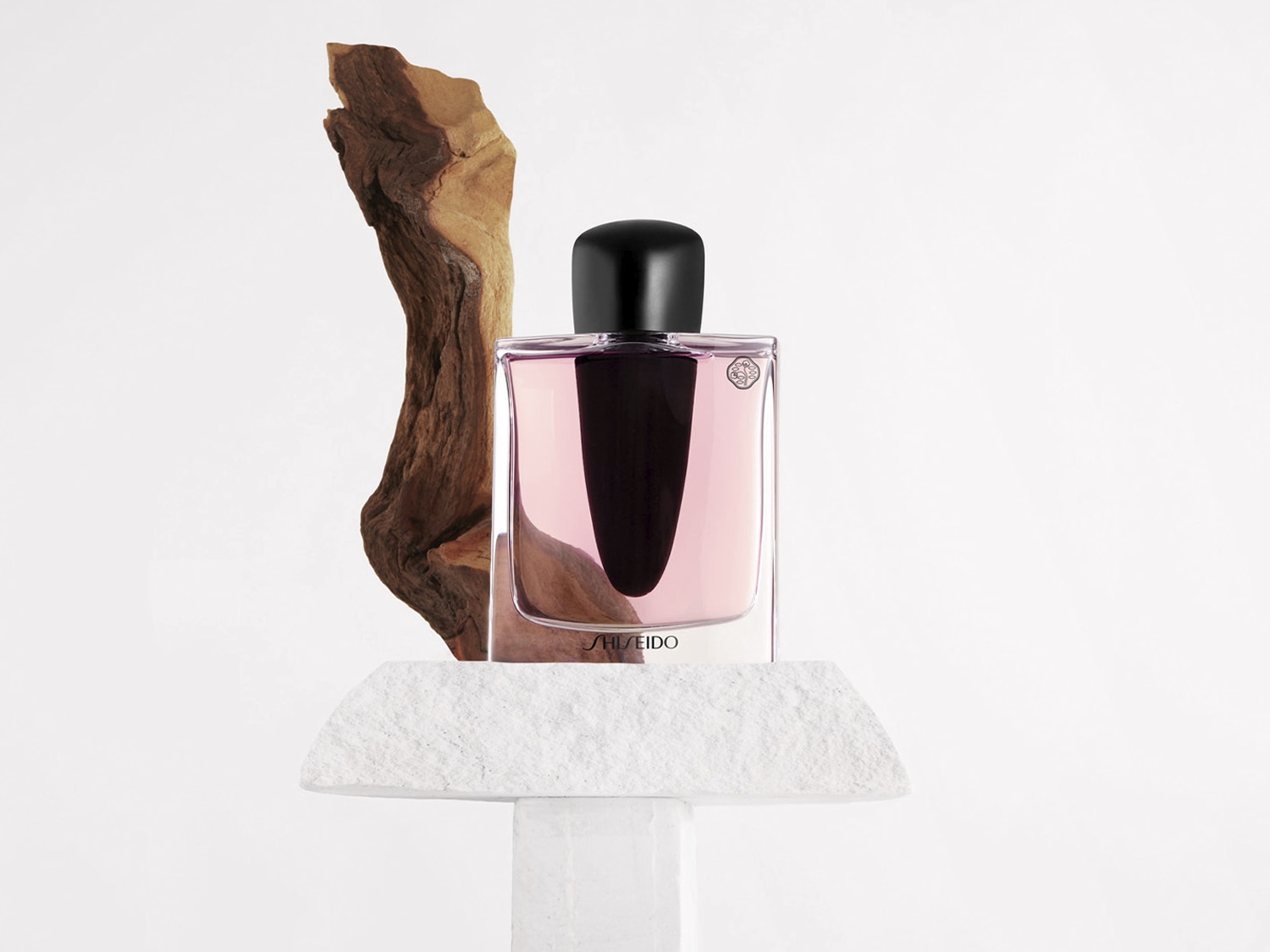 A Shiseido rózsaszín parfümös üvege egy fehér kőtalapzaton áll, mellette egy darab faág látható.