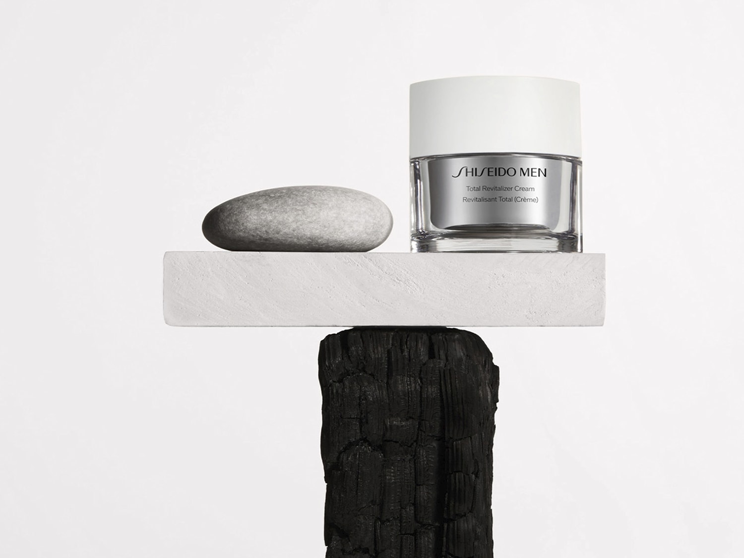 A Shiseido Men Total Revitalizer Cream egy átlátszó üveg tégelyben, ezüst színű kupakkal, egy szürke kő mellett egy fehér platformon helyezkedik el, amely egy elszenesedett fa tetején áll.