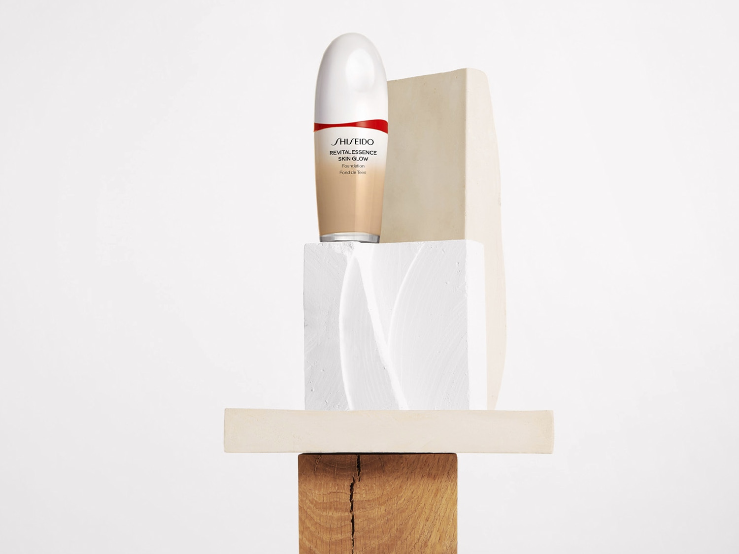 A Shiseido Revitalessence Skin Glow alapozó bézs színű, fehér kupakkal és piros csíkkal, geometrikus formákból álló tornyon áll.