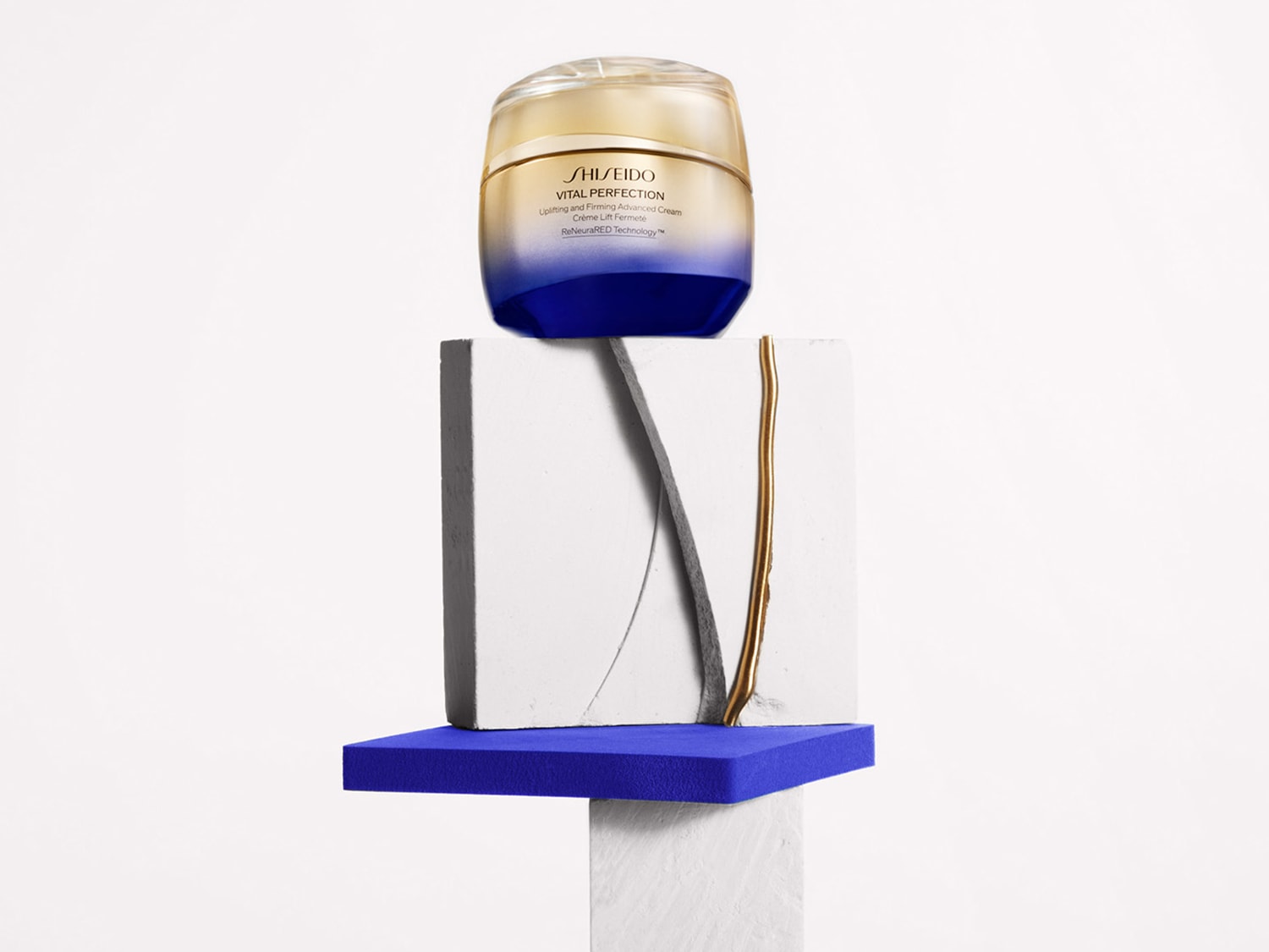 A Shiseido Vital Perfection krém elegáns, arany és kék tégelyben, amely egyedi, fehér és kék geometriai formákon helyezkedik el.