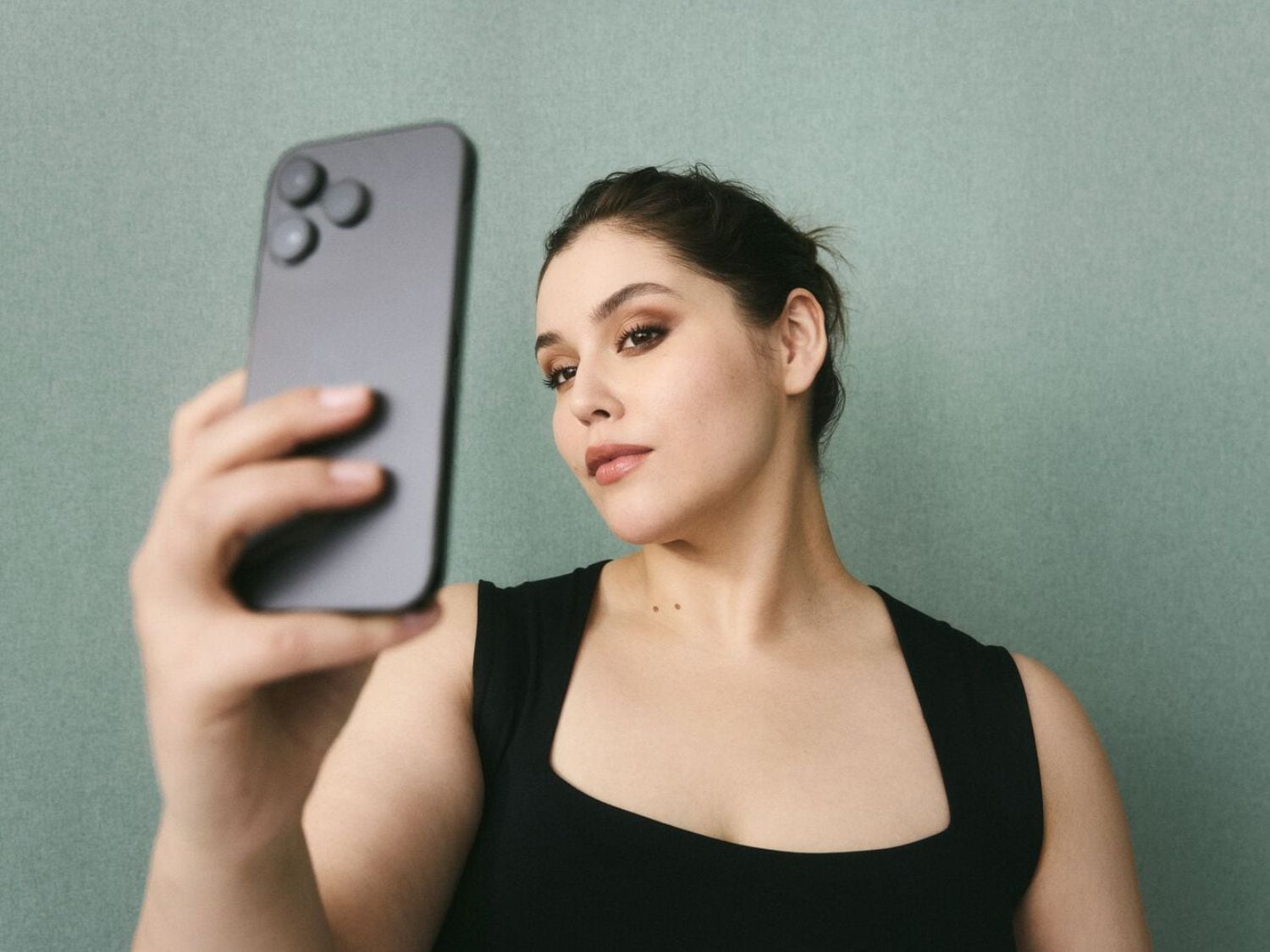 Une femme prend un selfie avec un smartphone gris, portant un débardeur noir et un maquillage subtil.