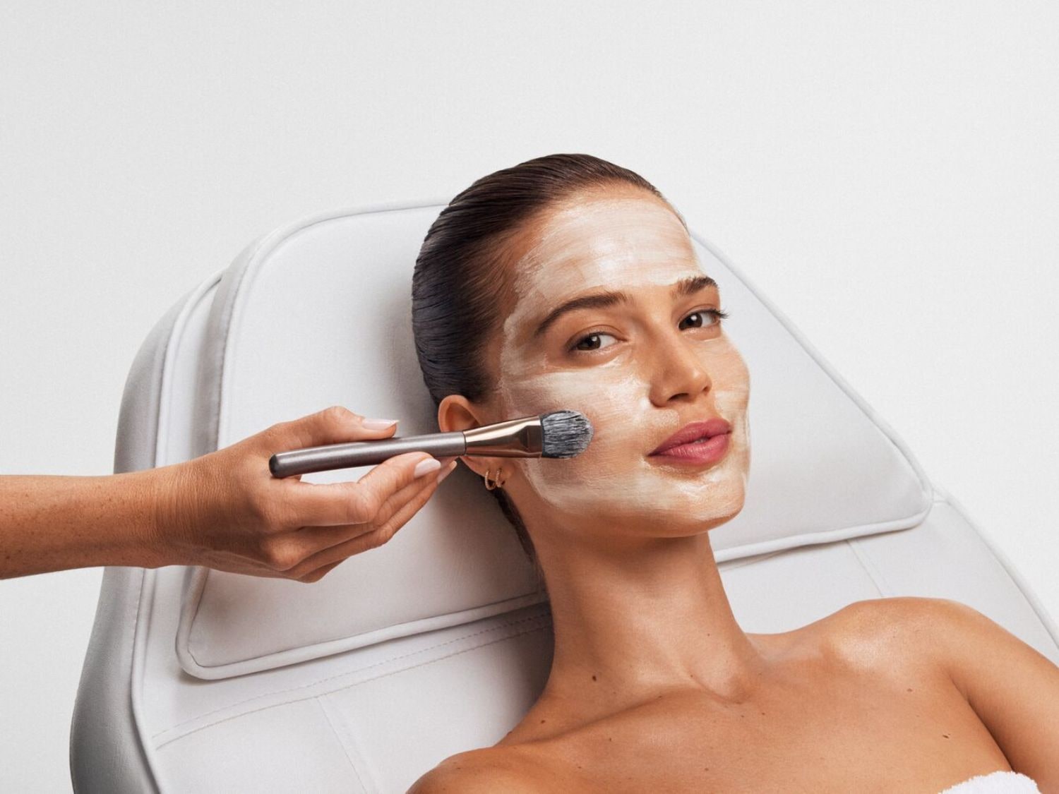 Une femme est allongée sur une chaise, un masque blanc lui est appliqué sur le visage avec un pinceau, pour un soin de beauté.