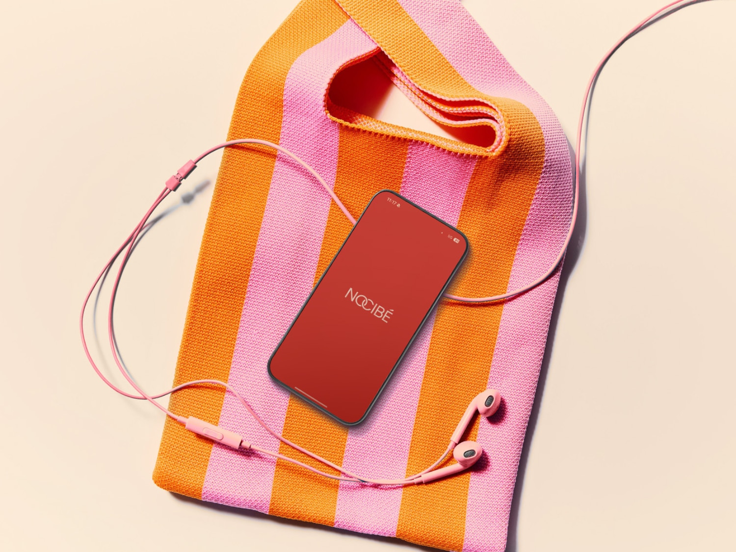Un sac en tricot rayé rose et orange avec un smartphone affichant le logo Nocibé et des écouteurs roses, le tout posé sur une surface claire.