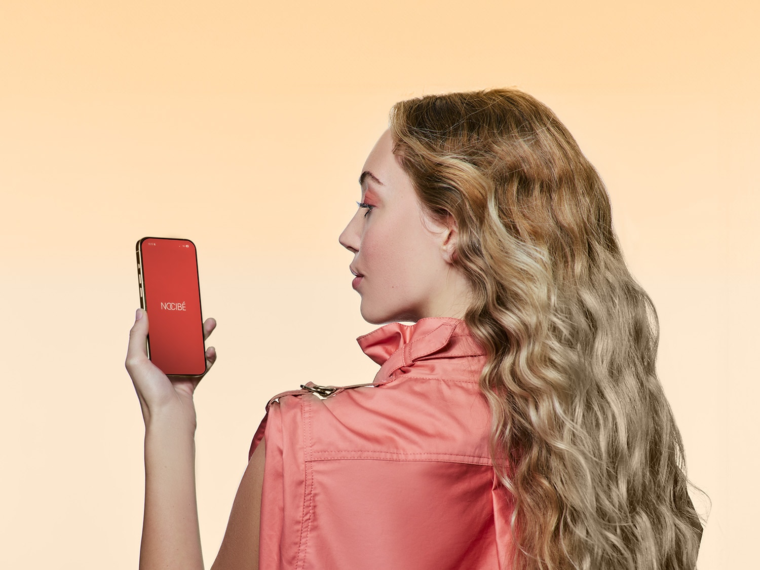 Une femme blonde regarde un smartphone rouge avec le logo Nocibé, portant une veste rose.