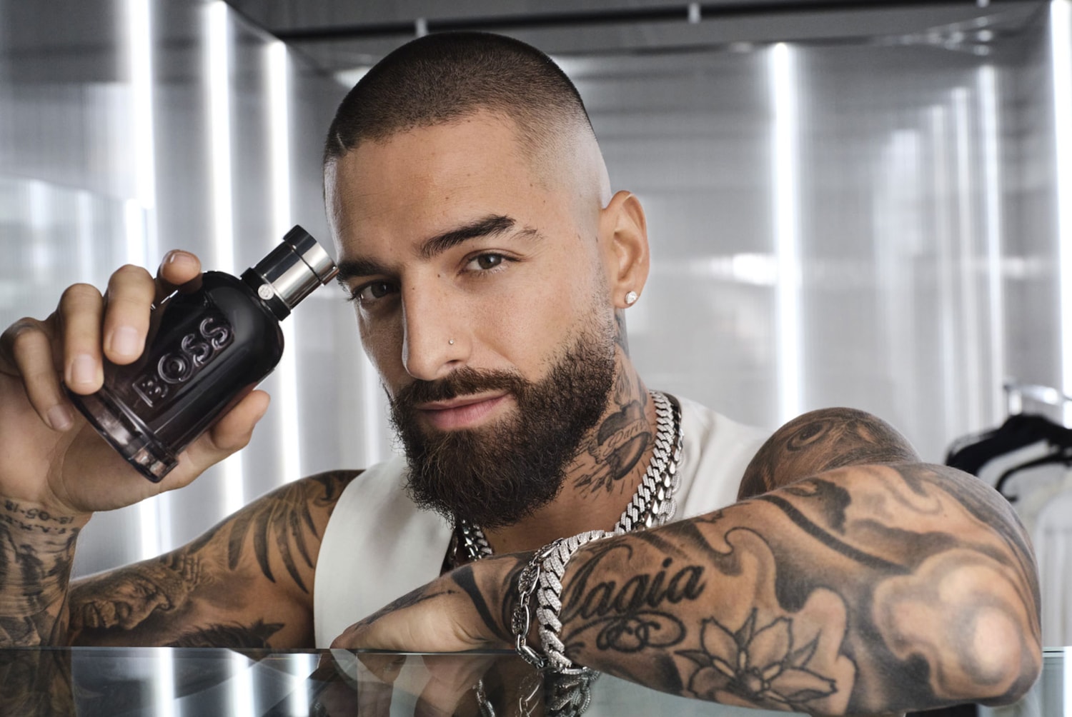 Maluma, tetoválásokkal és ezüst nyaklánccal, egy sötét BOSS parfümös üveget tart a kezében, miközben a kamerába néz.