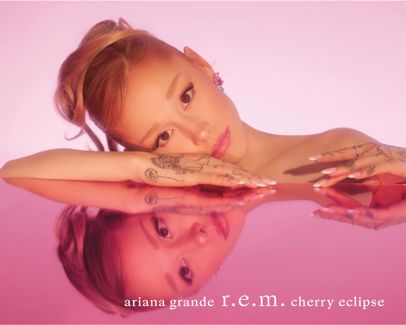 Na różowym tle Ariana Grande prezentuje R.E.M. Cherry Eclipse, opierając się o błyszczącą powierzchnię, która odbija jej twarz.