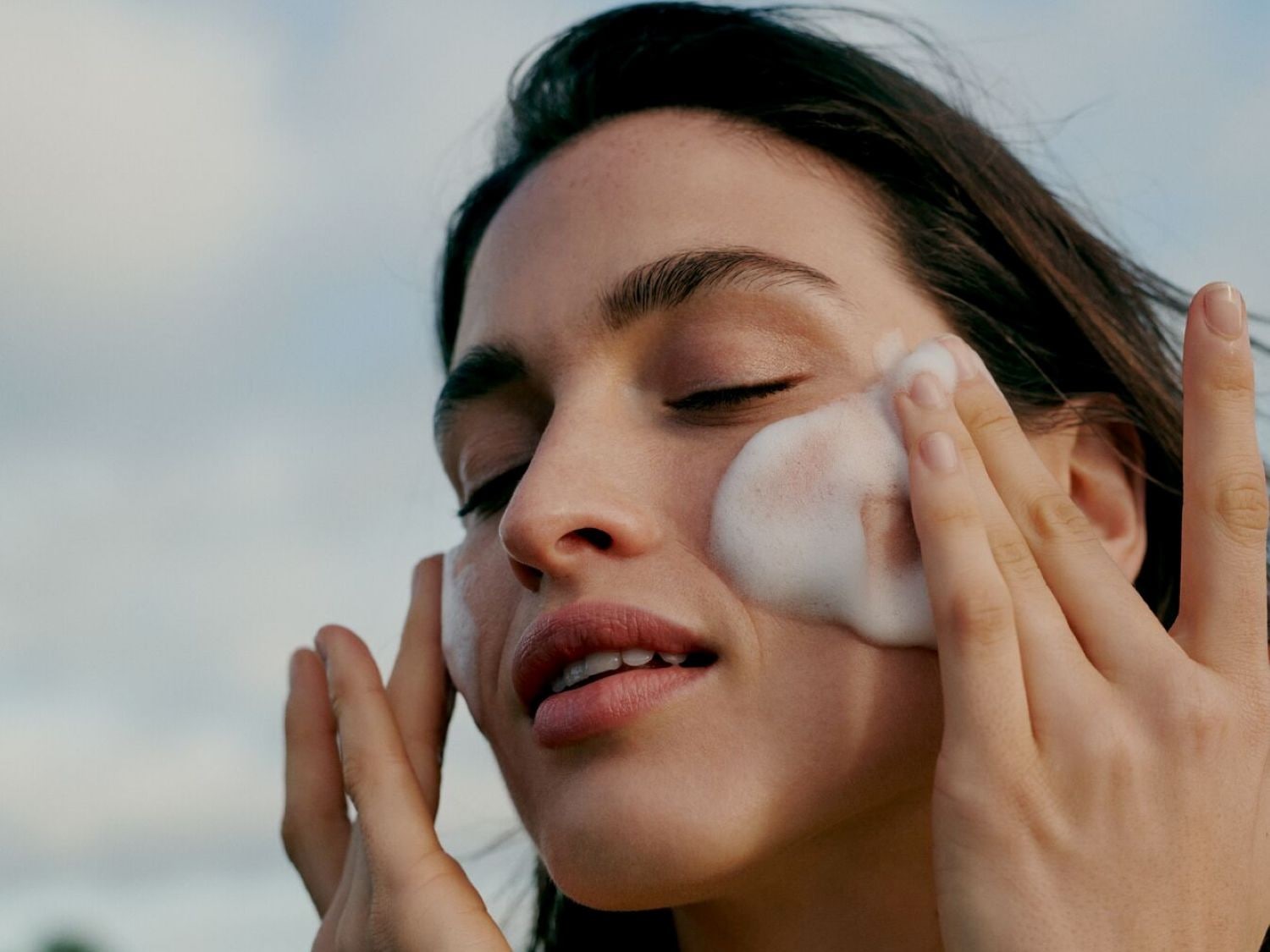Una donna con gli occhi chiusi si massaggia il viso con una schiuma detergente bianca, per una pelle pulita e fresca.