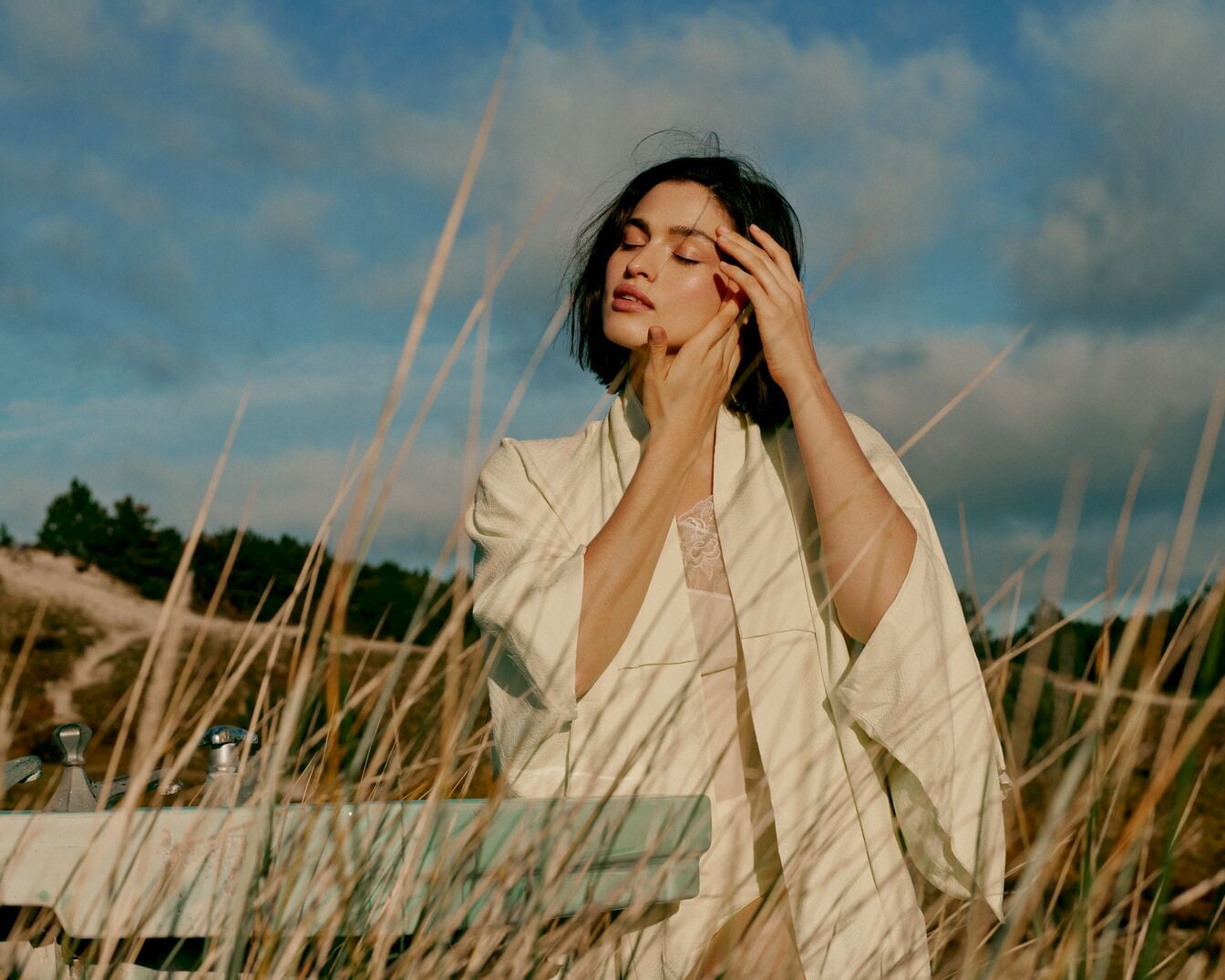 Una donna con indosso un kimono color crema chiaro, in piedi tra l'erba alta, con le mani che le toccano delicatamente il viso, gli occhi chiusi e un cielo azzurro sullo sfondo.