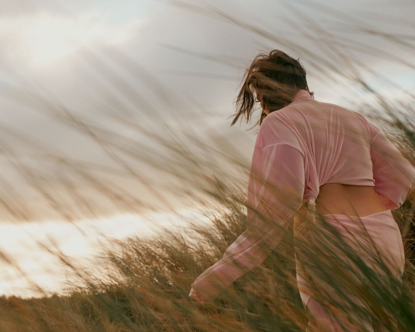Una donna con indosso un abito rosa chiaro cammina tra l'erba alta, con la schiena parzialmente scoperta e i capelli mossi dal vento.