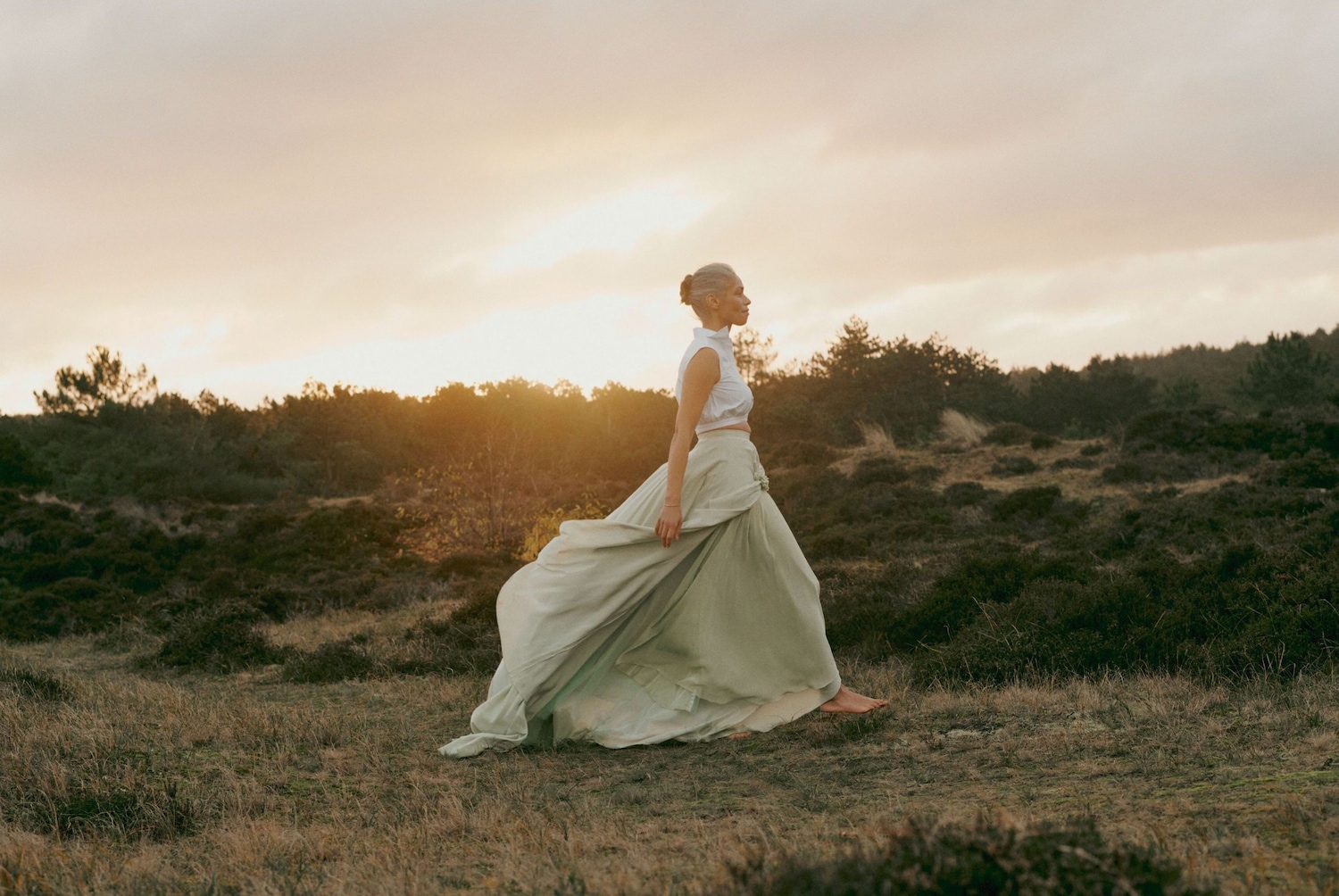 Una donna con un abito fluente color verde pallido cammina a piedi nudi in un campo al tramonto, incarnando un'eleganza eterea e un legame con la natura.