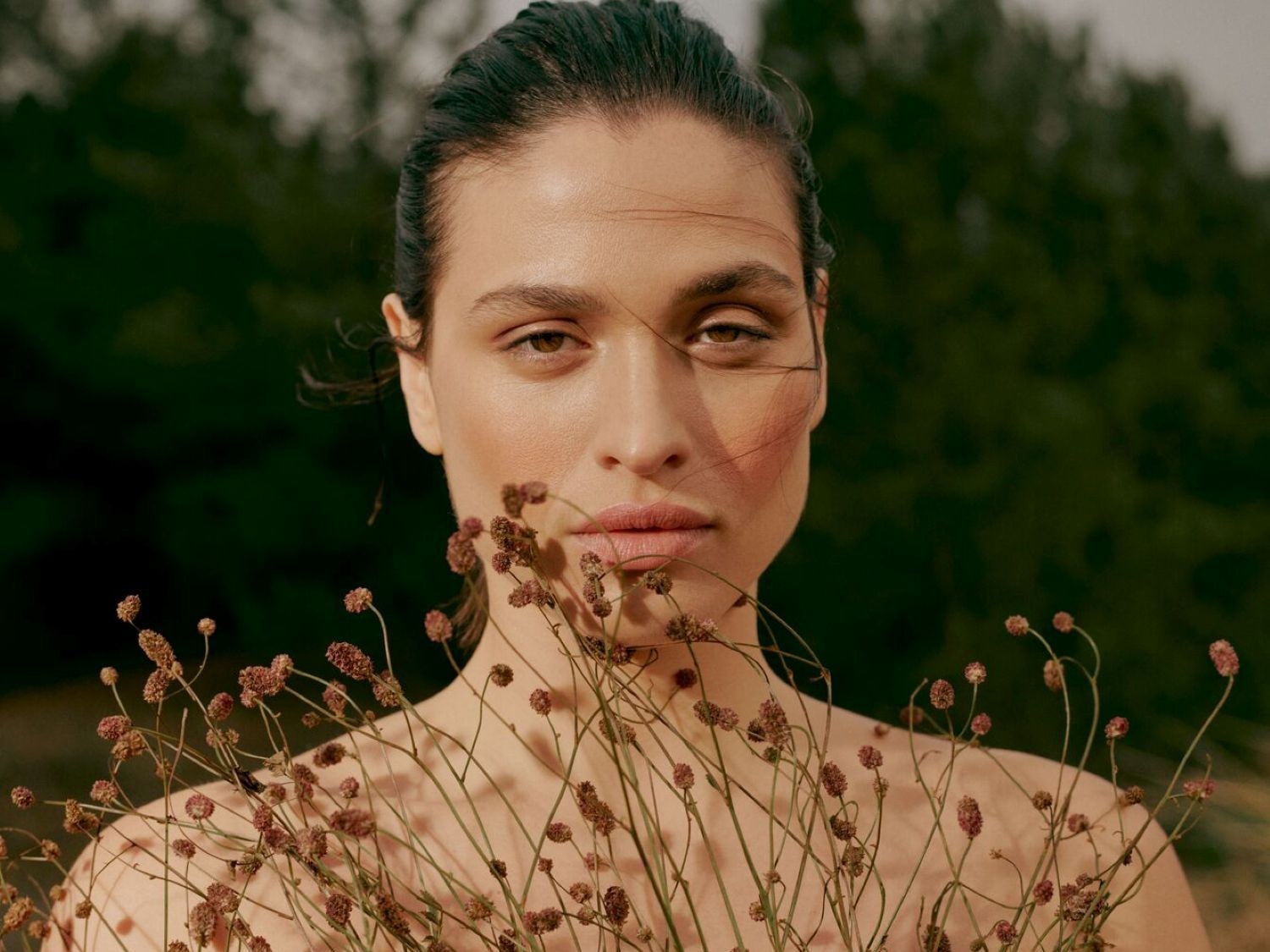 Primo piano di una donna con capelli scuri tirati indietro, pelle luminosa e un delicato trucco naturale, che tiene un mazzetto di fiori secchi davanti a uno sfondo verde scuro.