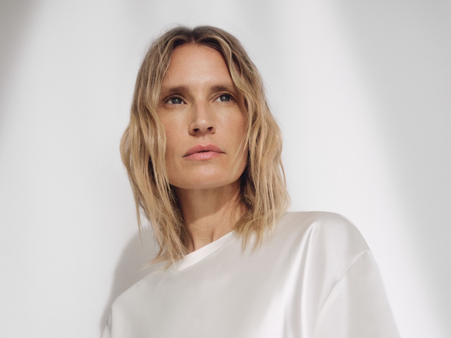 Een vrouw met blond haar en een witte satijnen blouse kijkt naar de verte, haar blik vol verwachting.