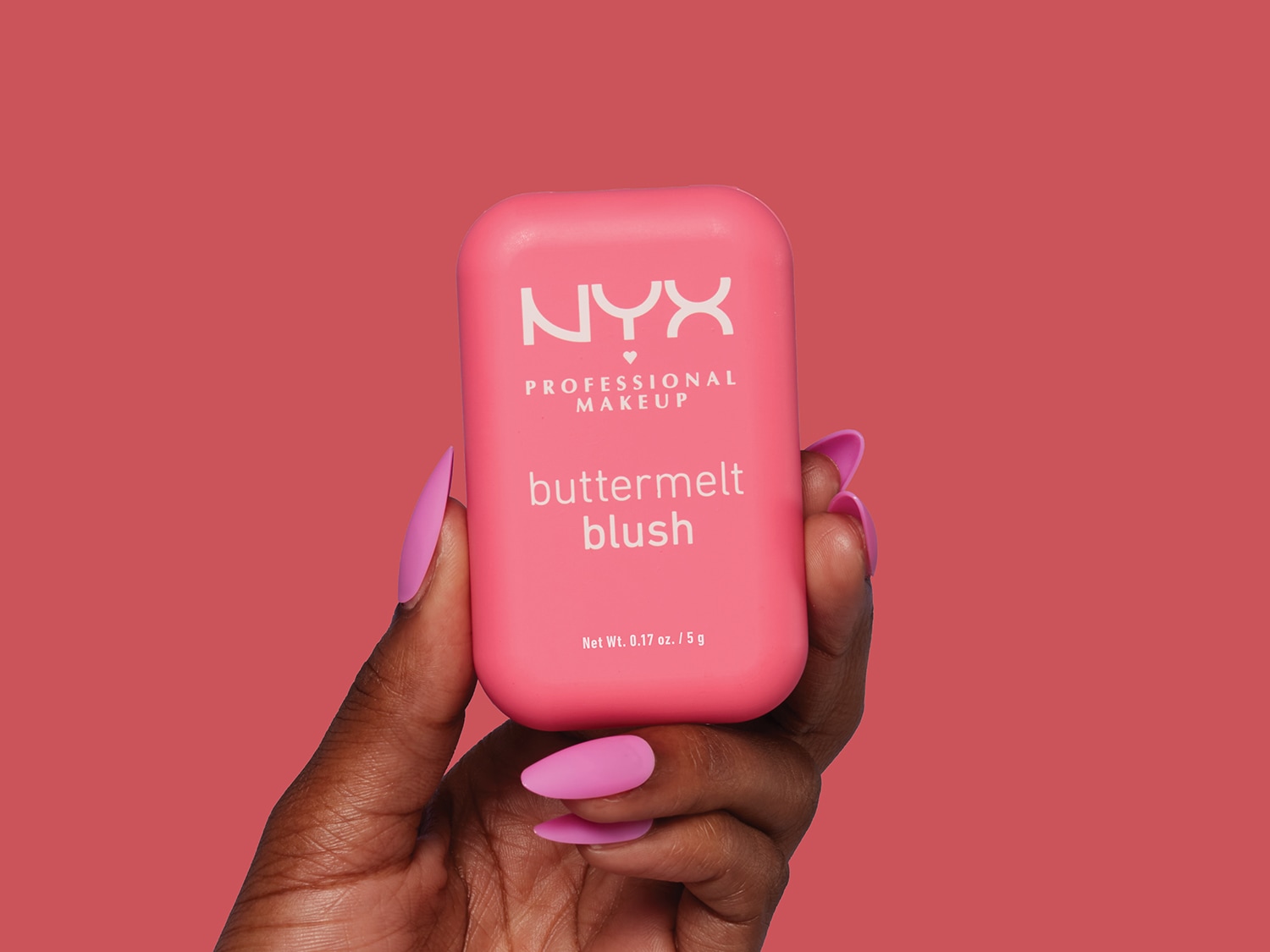 Une main tenant un blush NYX Professional Makeup Buttermelt de couleur rose, avec un fond rose assorti.