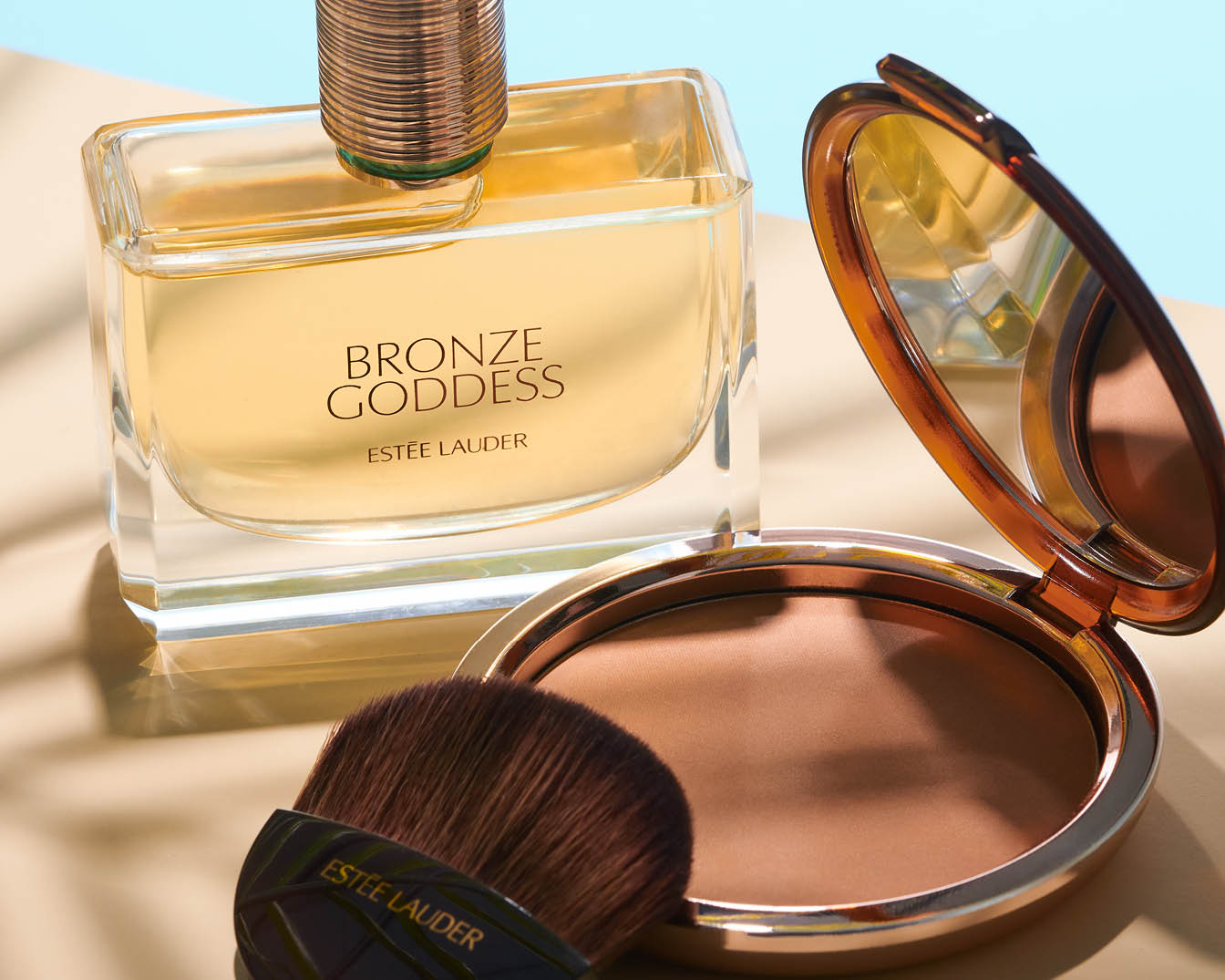 Découvrez le parfum Bronze Goddess d'Estée Lauder dans son flacon en verre transparent, accompagné d'une poudre bronzante et d'un pinceau pour un teint ensoleillé.