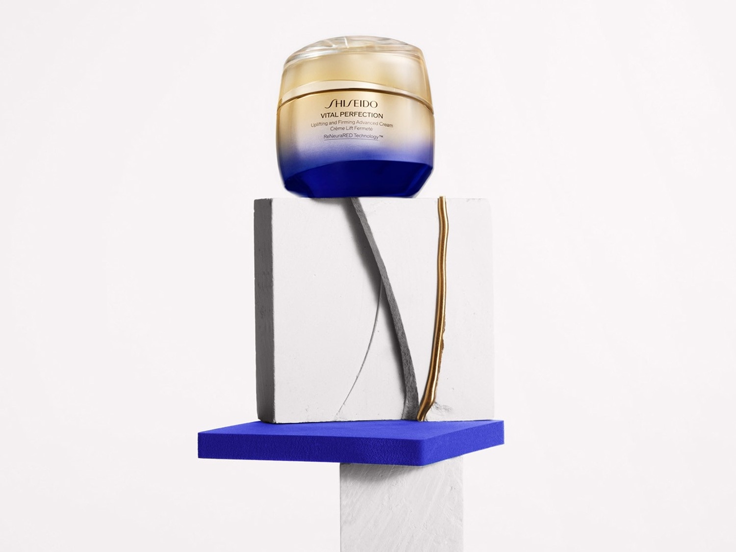 Krem Shiseido Vital Perfection Uplifting and Firming Advanced Cream w słoiczku o złotym wieczku i granatowej podstawie, umieszczony na białym postumencie z niebieskimi akcentami.