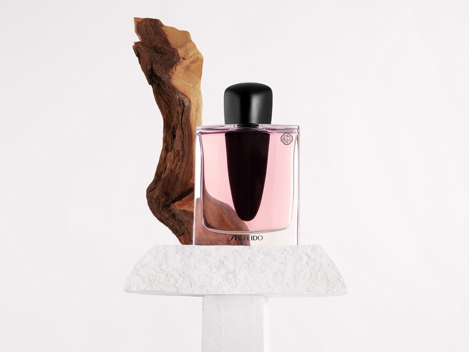 Perfumy Shiseido w przezroczystej butelce z różową zawartością i czarną zakrętką, stoją na białym kamieniu obok kawałka drewna.