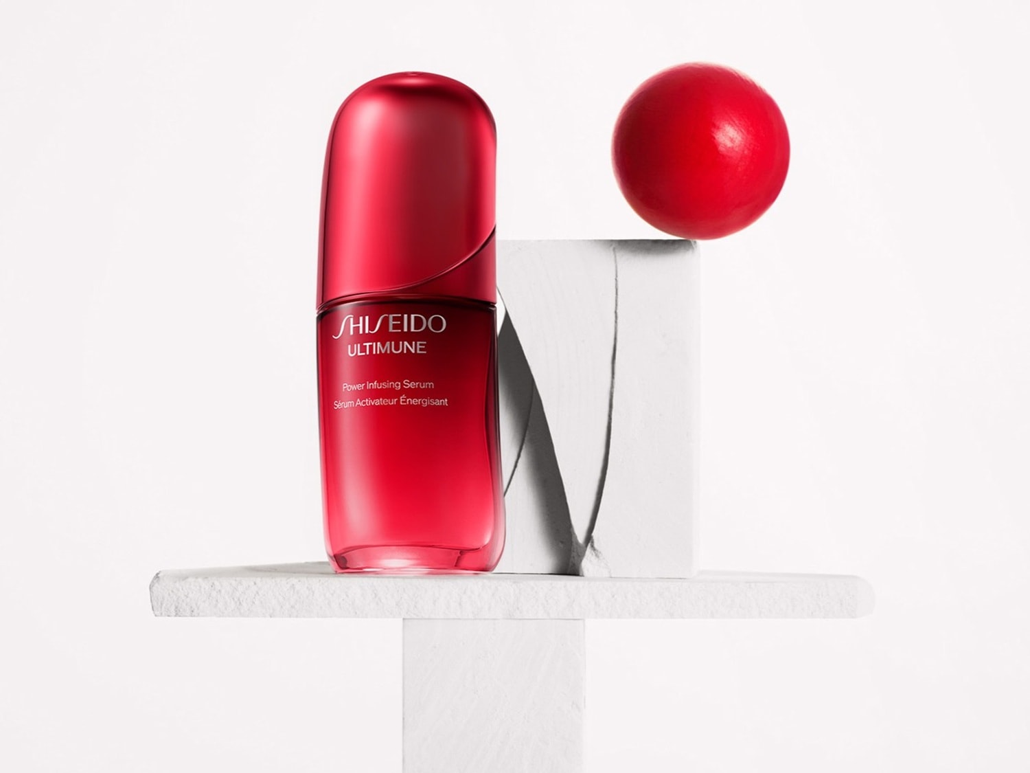 Czerwone serum Shiseido Ultimune Power Infusing Serum w szklanej butelce, stojące na białym, kamiennym postumencie z czerwoną kulką na górze, na białym tle.