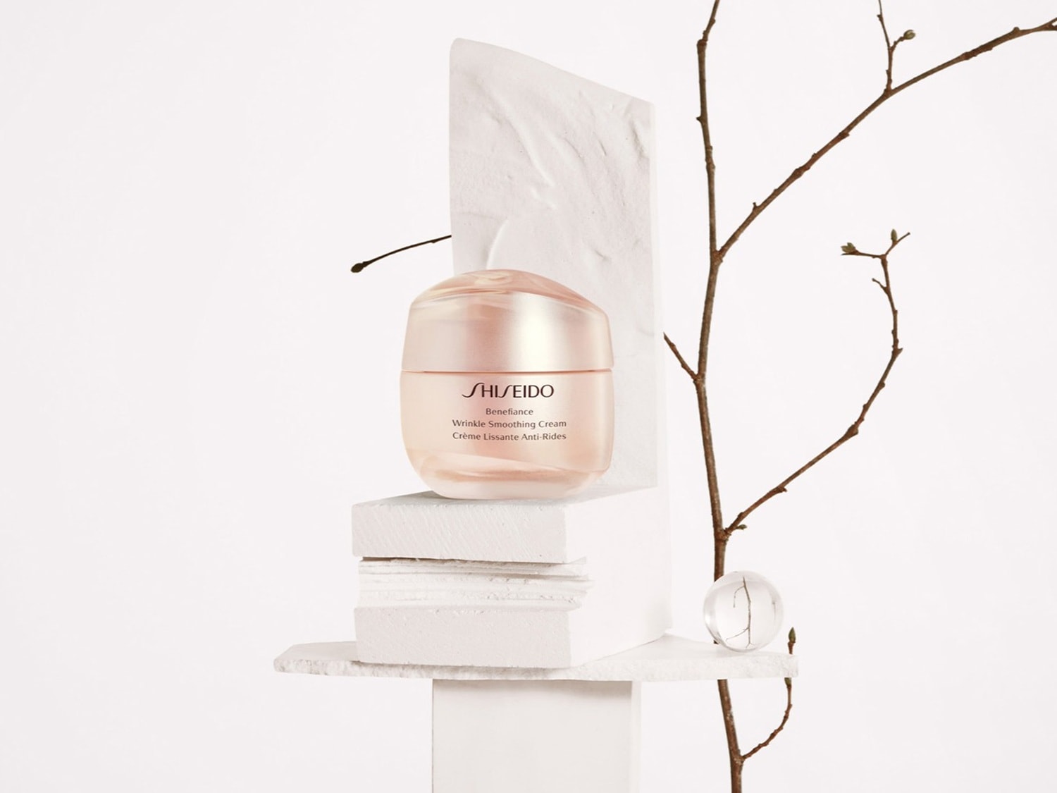 Krem Shiseido Benefiance Wrinkle Smoothing Cream w pudrowo-różowym słoiczku, ustawiony na białych kamieniach, obok gałązki, na białym tle.
