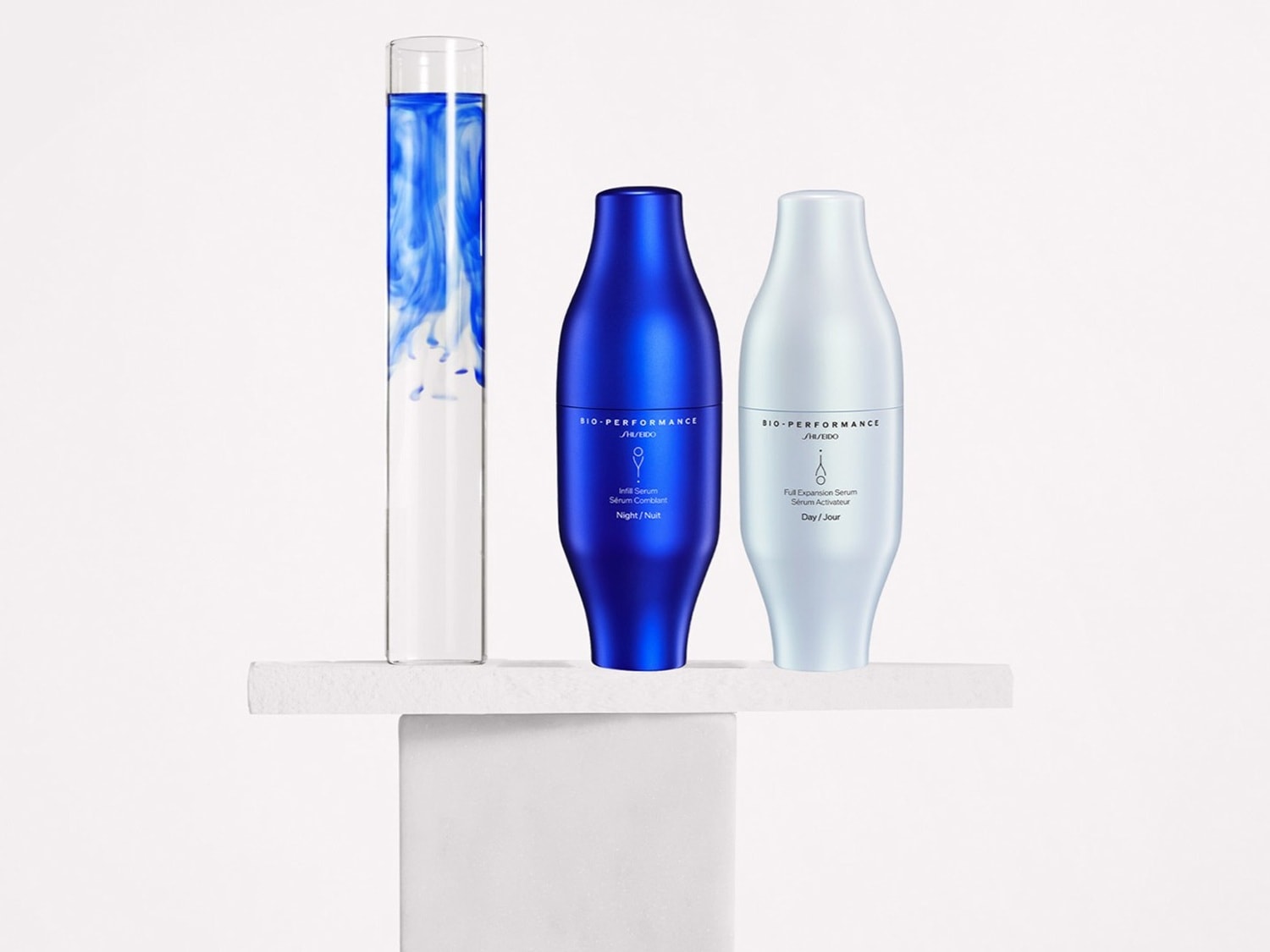 Na białym tle, na podeście, znajdują się dwa flakony Shiseido Bio-Performance: granatowy Infill Serum na noc i srebrny Full Expansion Serum na dzień, obok nich szklana tuba z niebieskim płynem.