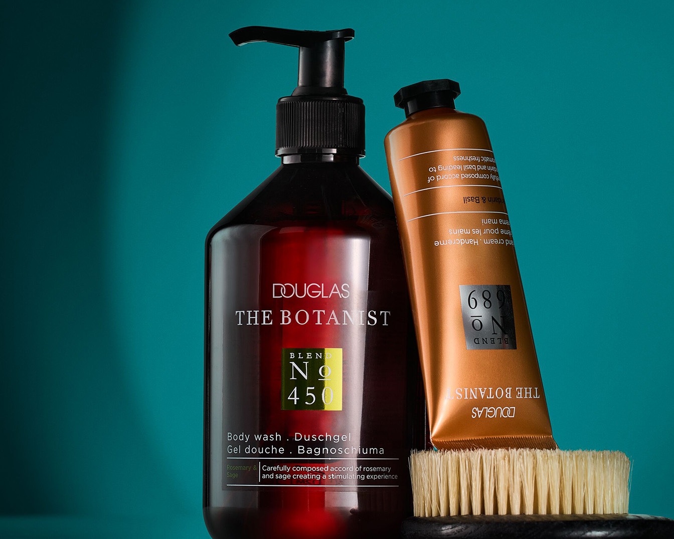 Dunkelrote Douglas The Botanist Duschgelflasche mit schwarzem Pumpspender, daneben eine goldene Handcremetube und eine Bürste auf blaugrünem Hintergrund.