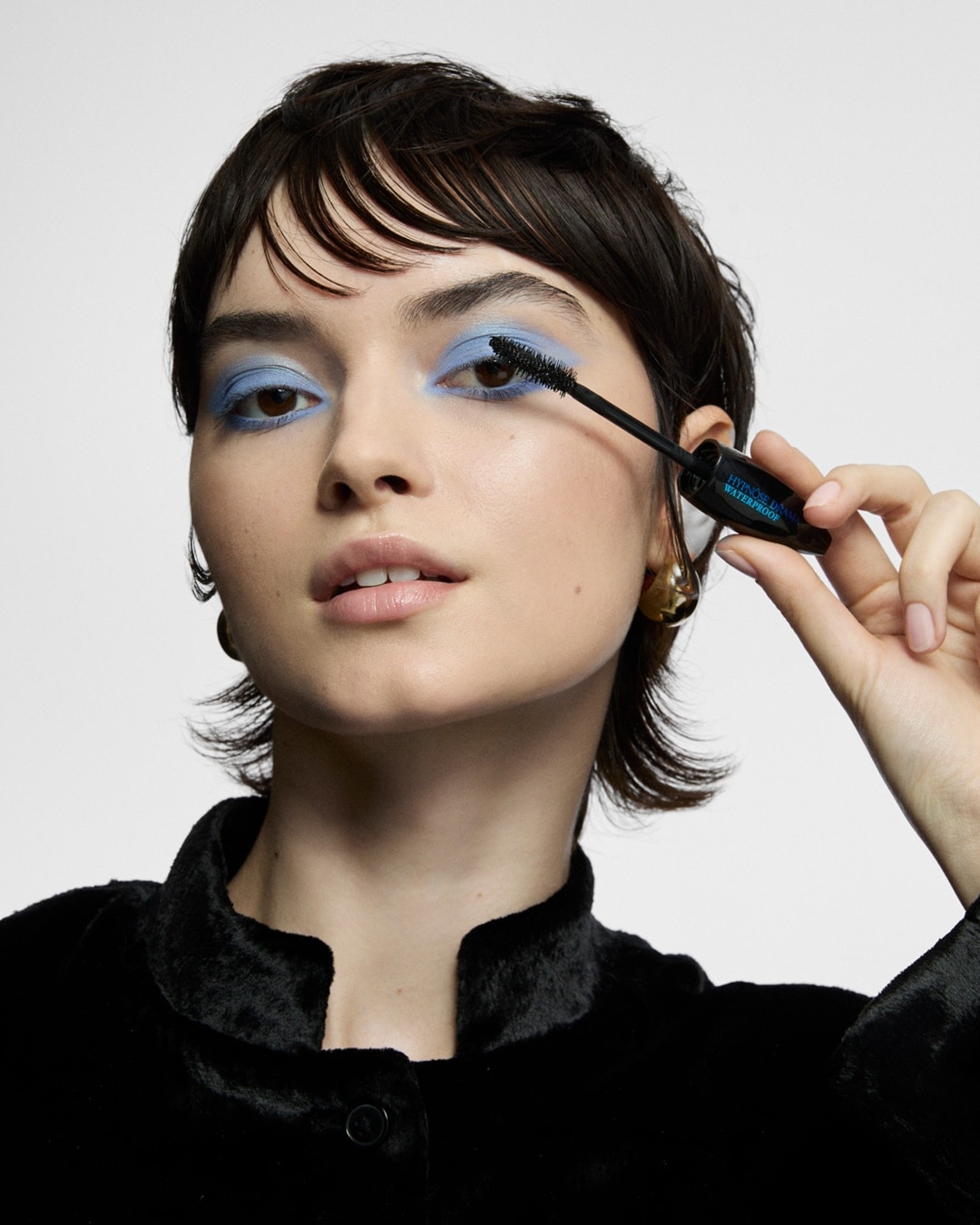 Eine Frau mit blauem Lidschatten trägt schwarze Wimperntusche mit einer Bürste auf ihre Wimpern auf.