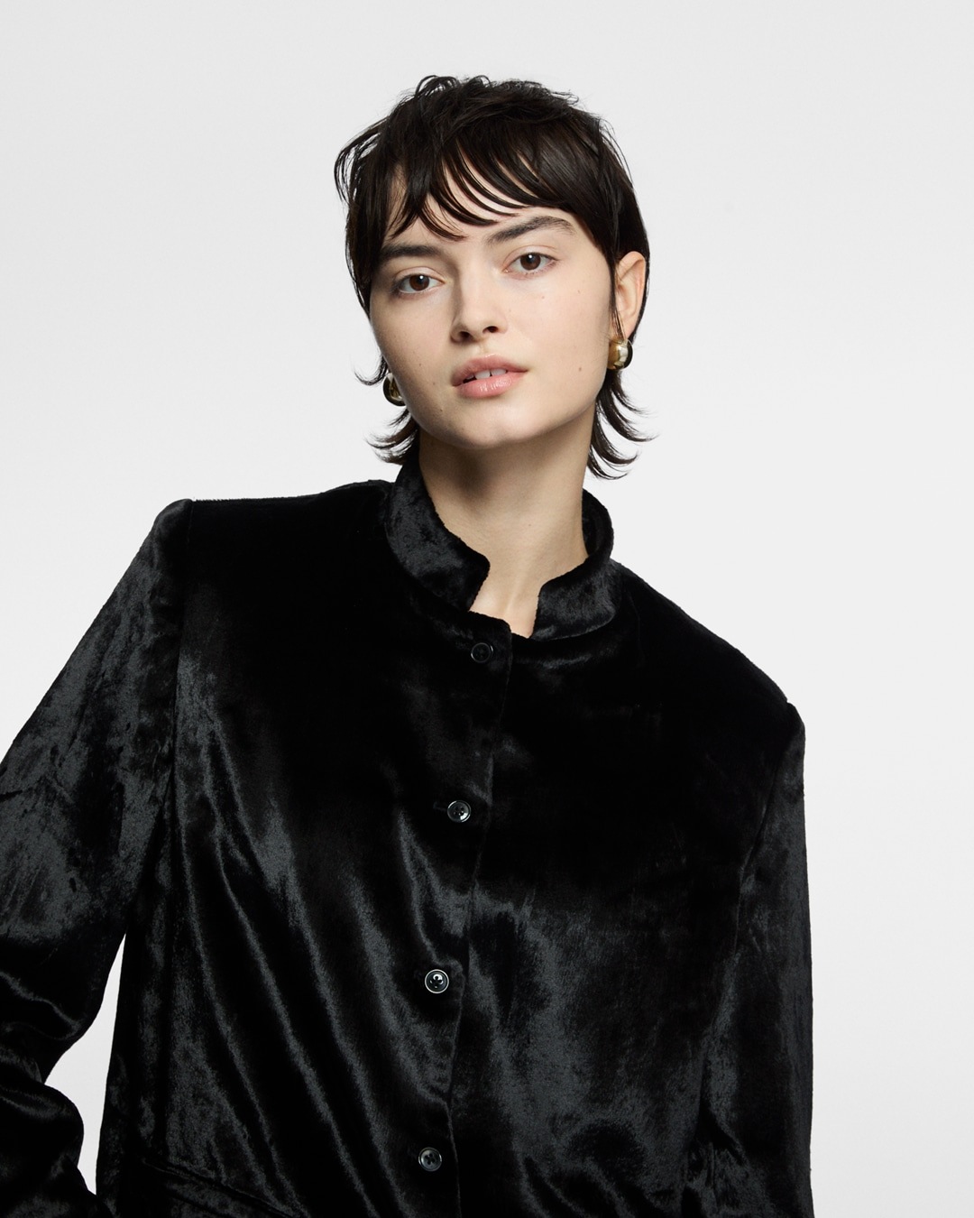 Eine Frau mit kurzen, dunklen Haaren trägt eine schwarze Samtjacke mit Stehkragen und goldenen Ohrringen vor einem neutralen Hintergrund.