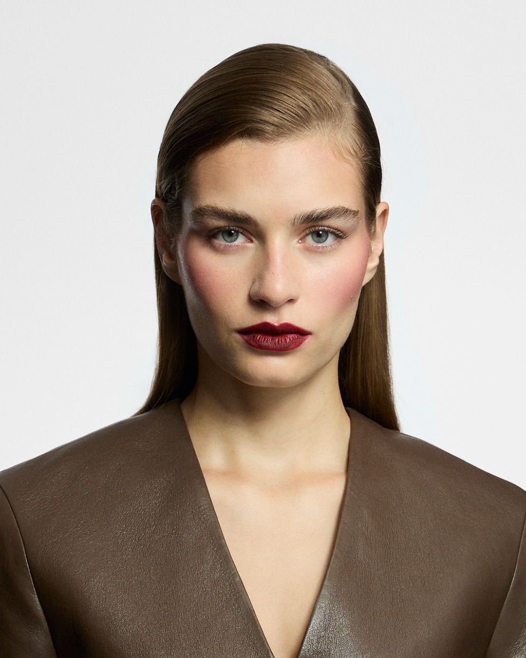 Porträt einer Frau mit glatt zurückgekämmtem, braunem Haar, die ein braunes Lederoberteil mit V-Ausschnitt trägt.