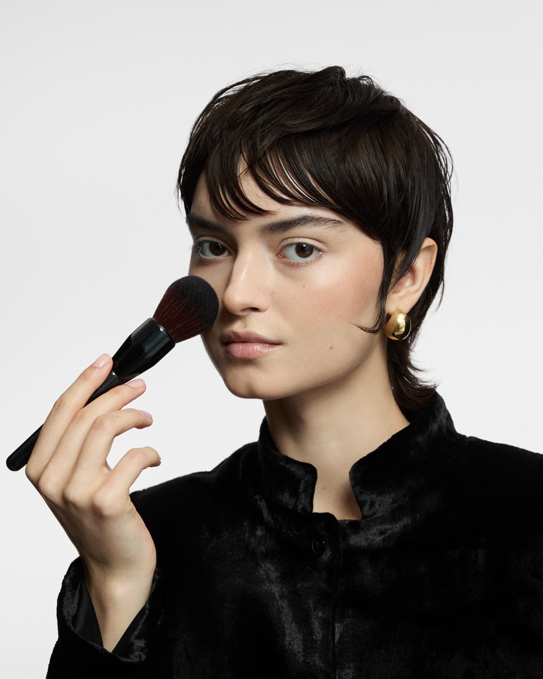 Eine Frau mit kurzen, dunklen Haaren und goldenen Ohrringen trägt mit einem Pinsel Make-up auf.