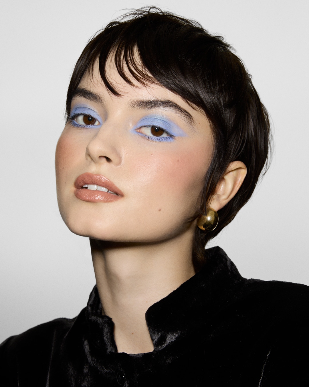 Eine Frau mit kurzen, dunklen Haaren und hellblauem Lidschatten, der ihre braunen Augen betont, trägt ein schwarzes Samtoberteil und goldene Ohrringe.